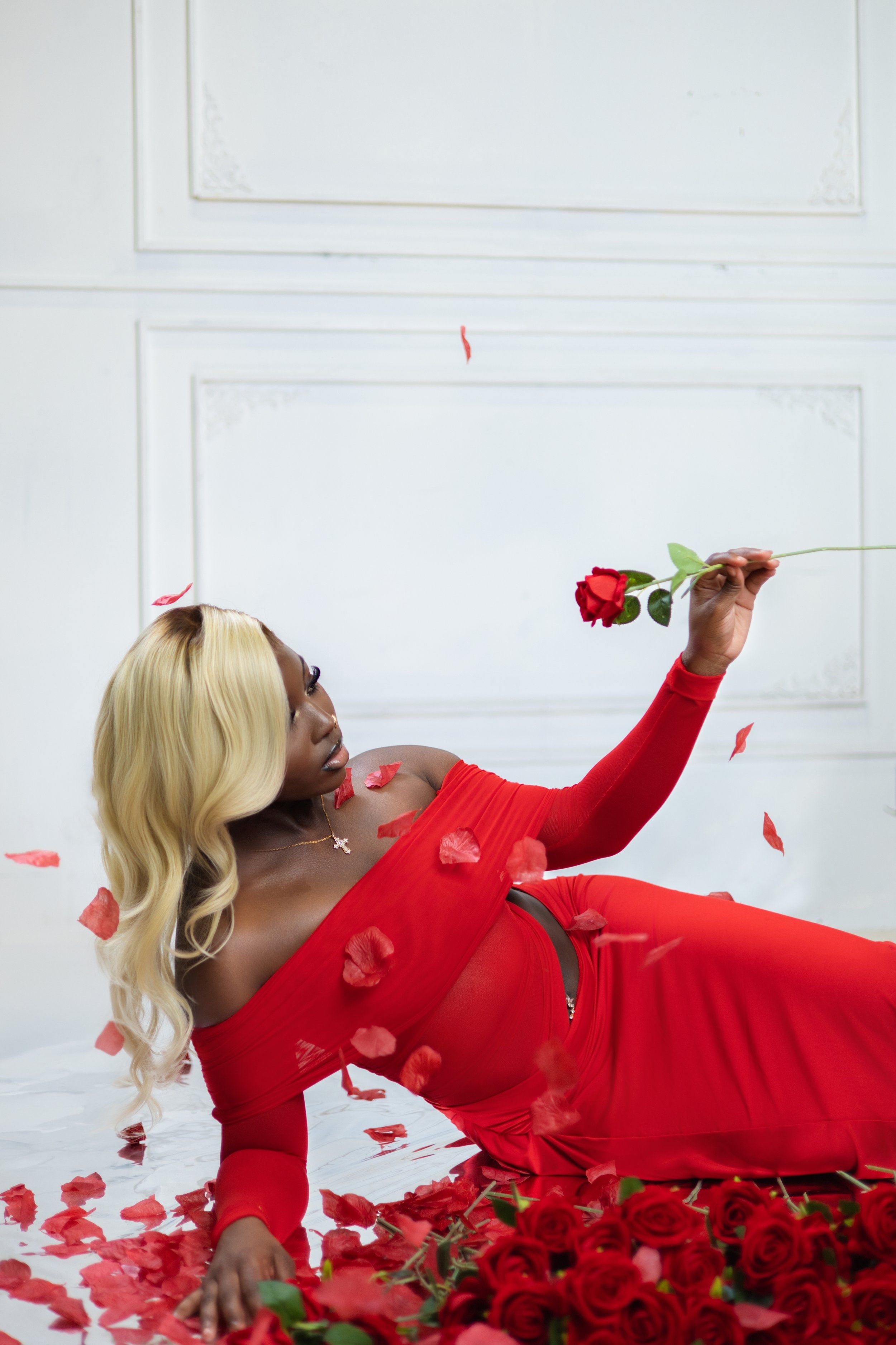 A woman with blonde hair in a red dress lying on the floor surrounded by red roses and rose petals, holding a single rose, with falling petals around her.