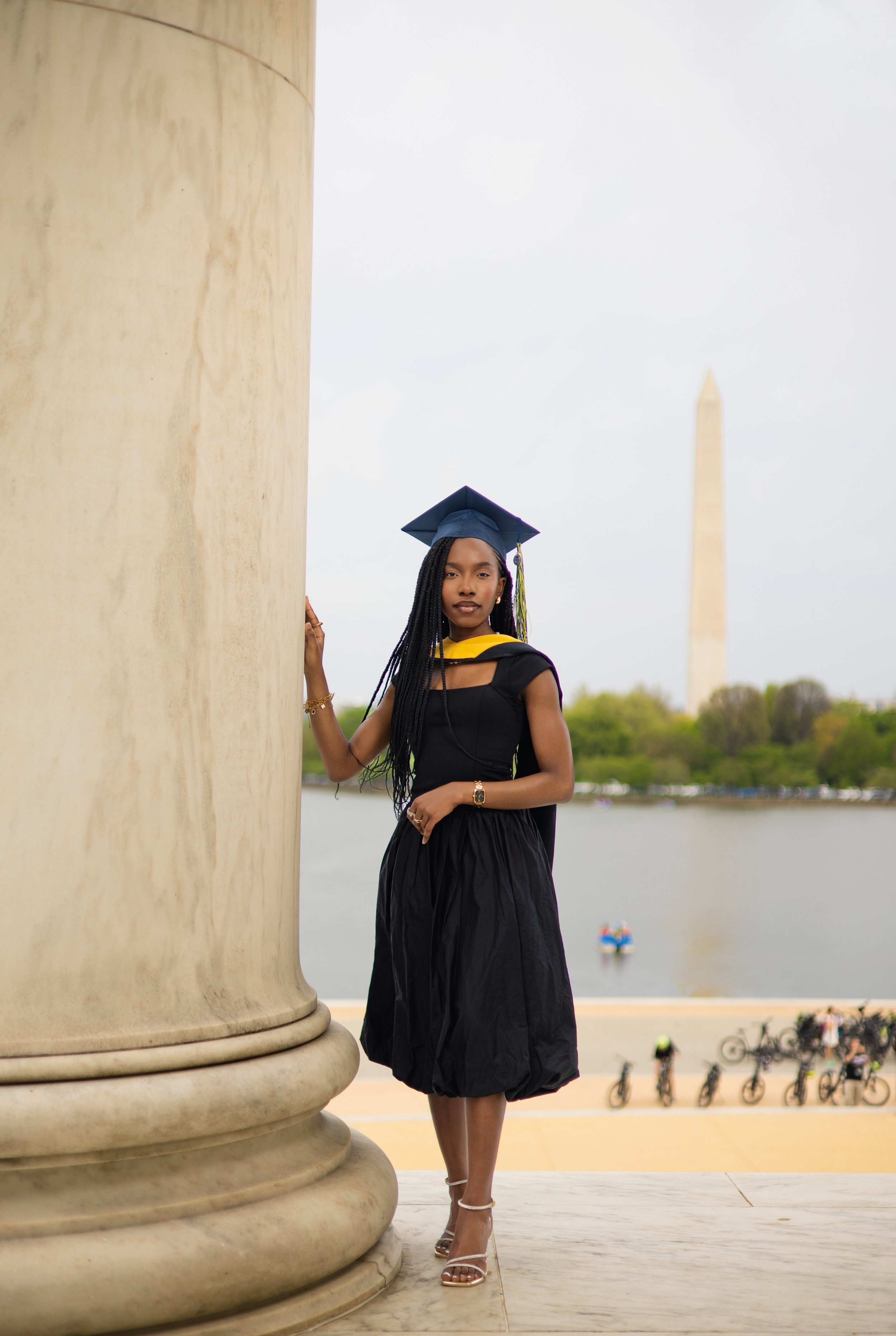 Young woman in black graduation gown and cap standing near a large stone column with the Washington Monument and water in the background. She is wearing high heels, a gold bracelet, and a watch, with a yellow stole around her shoulders.