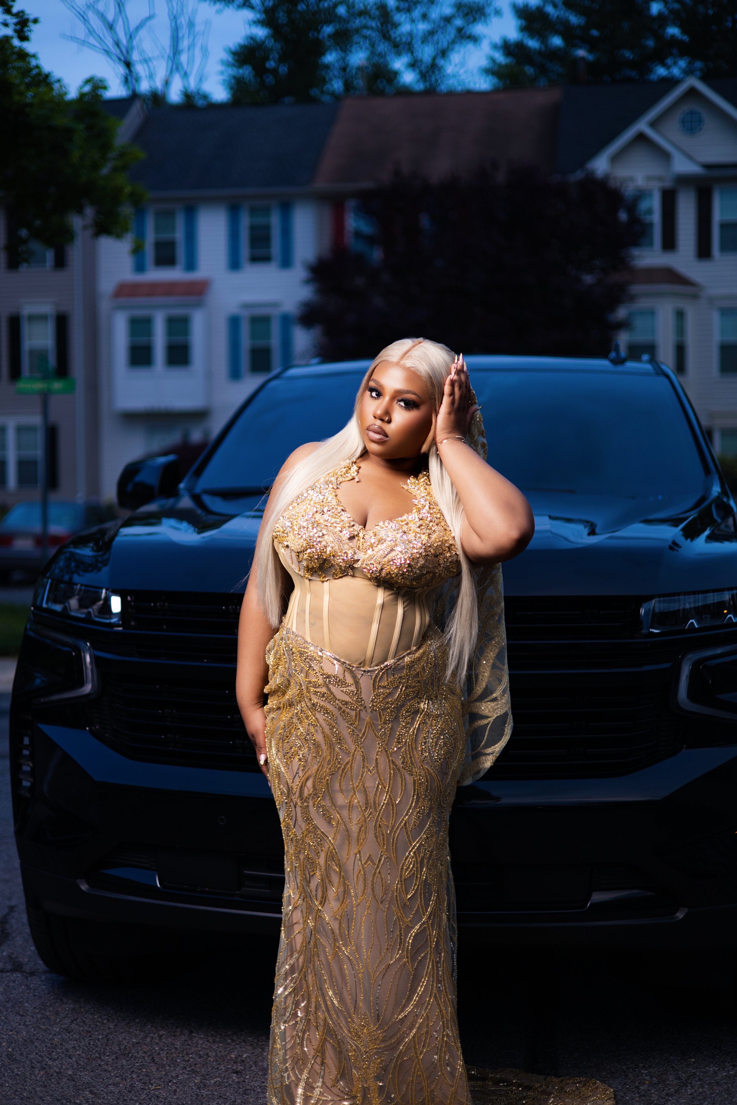 A woman with long platinum blonde hair wearing a gold, intricately embroidered gown poses in front of a black car on a residential street during dusk.