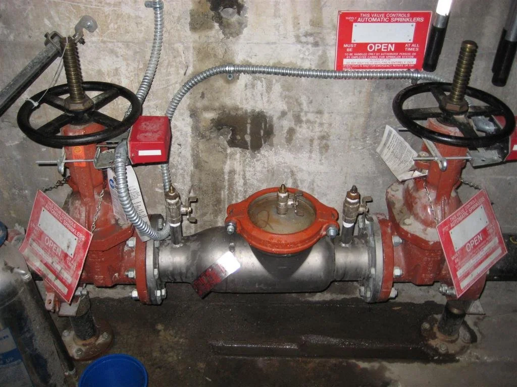 A fire sprinkler control valve with two red wheel handles, a central valve, and warning tags, installed on a concrete wall.