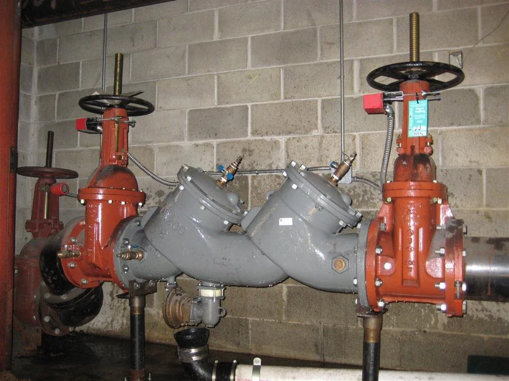 Industrial pipes and valves mounted on a concrete wall, with a gray Y-shaped pipe connection in the center and red valve handles on top.