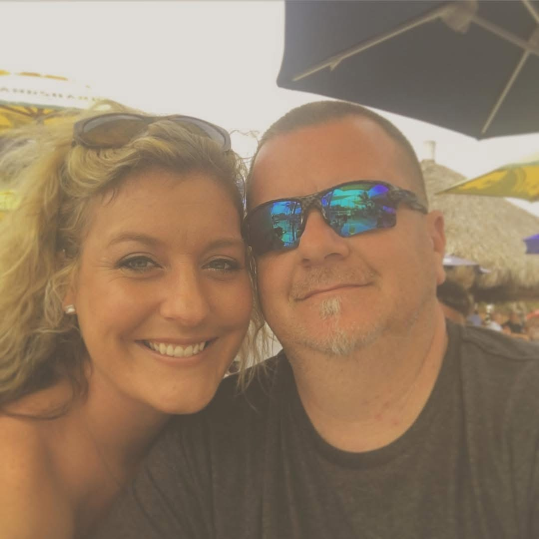 A smiling woman and a man wearing sunglasses take a close-up selfie outdoors, with umbrellas and a thatched roof in the background.