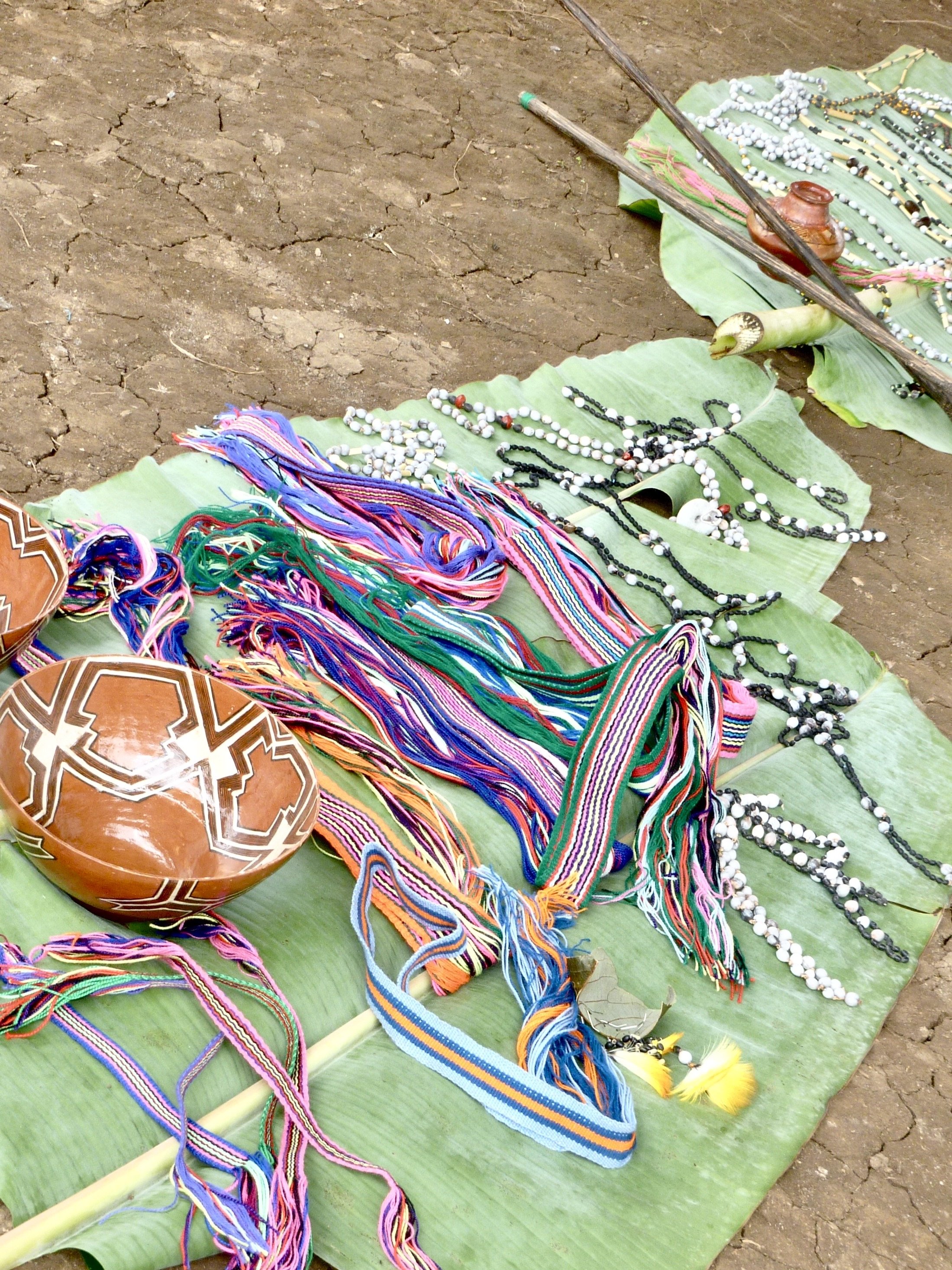 Colorful handmade necklaces and bracelets laid out on green banana leaves on dry cracked earth.