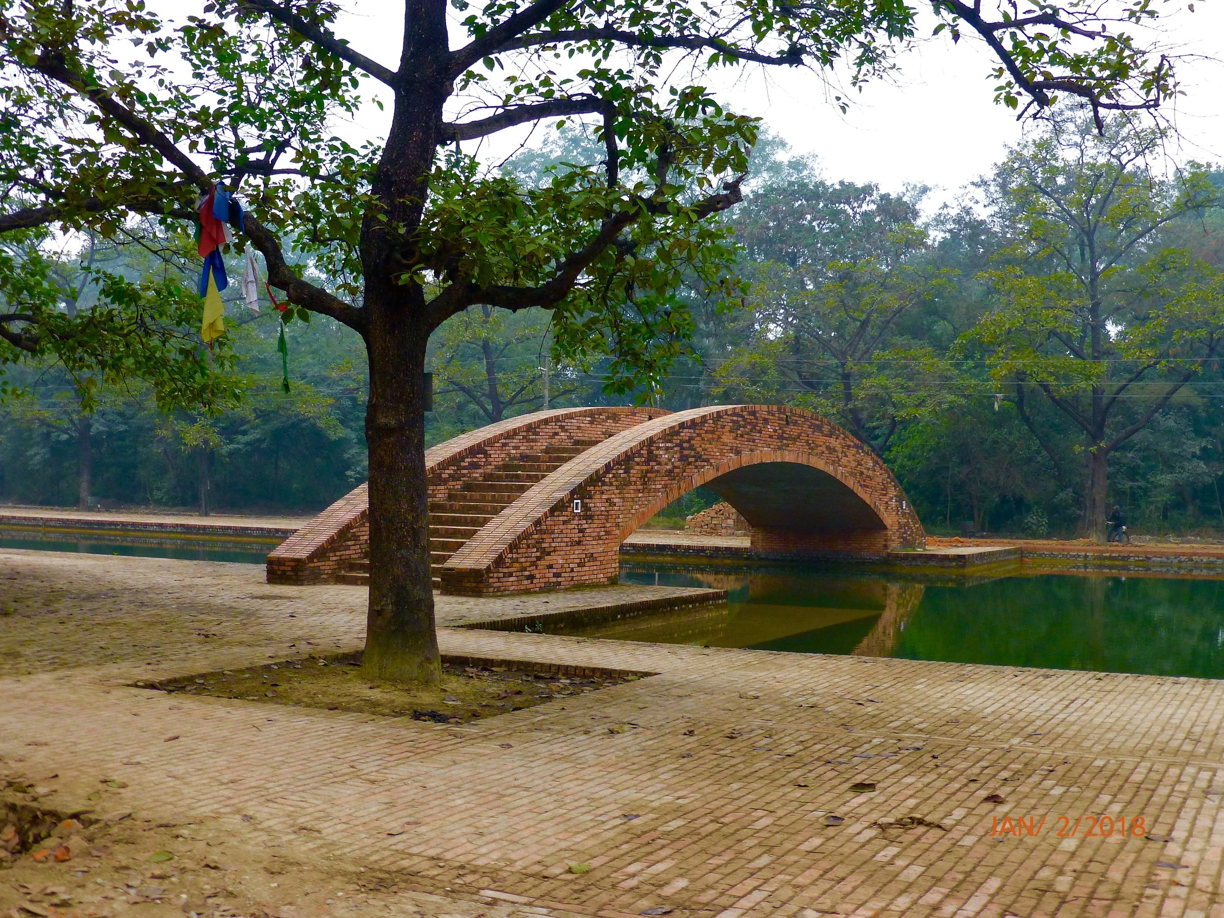 A brick bridge over a pond in a park with trees and greenery, and a person riding a bicycle in the background. The scene appears misty or foggy.