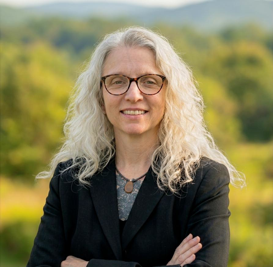 A woman with long, curly gray hair and glasses smiling outdoors with a background of green trees and a cloudy sky.