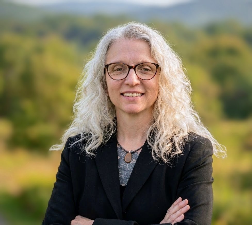 A woman with long, curly gray hair and glasses smiling outdoors, with a blurred green landscape in the background.