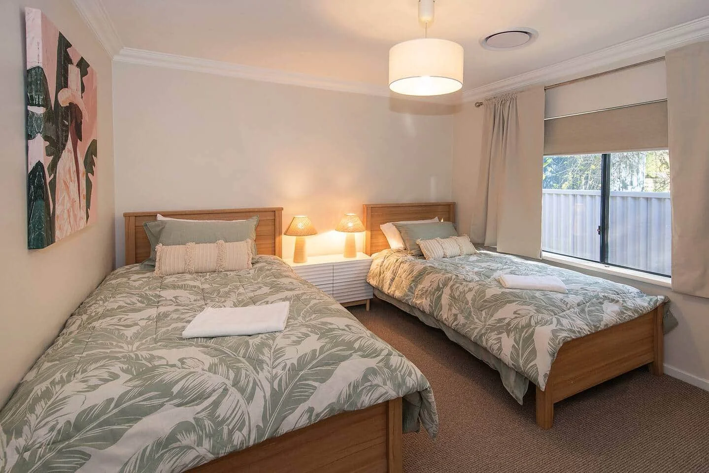 Bedroom with two beds, floral-patterned bedding, wooden headboards, a window with curtains, a painting on the wall, and a nightstand with two lamps.