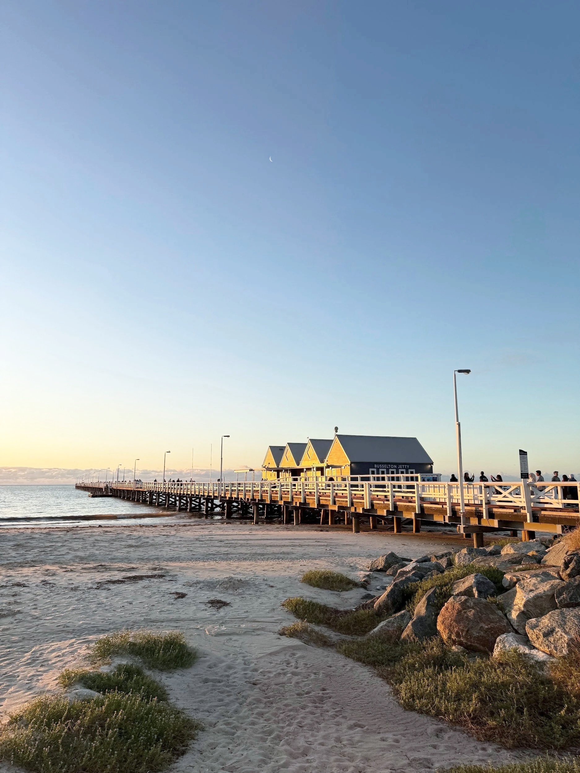 A wooden pier extends into the ocean with small beach huts on the end, under a clear sky with a visible crescent moon, during sunset or sunrise.