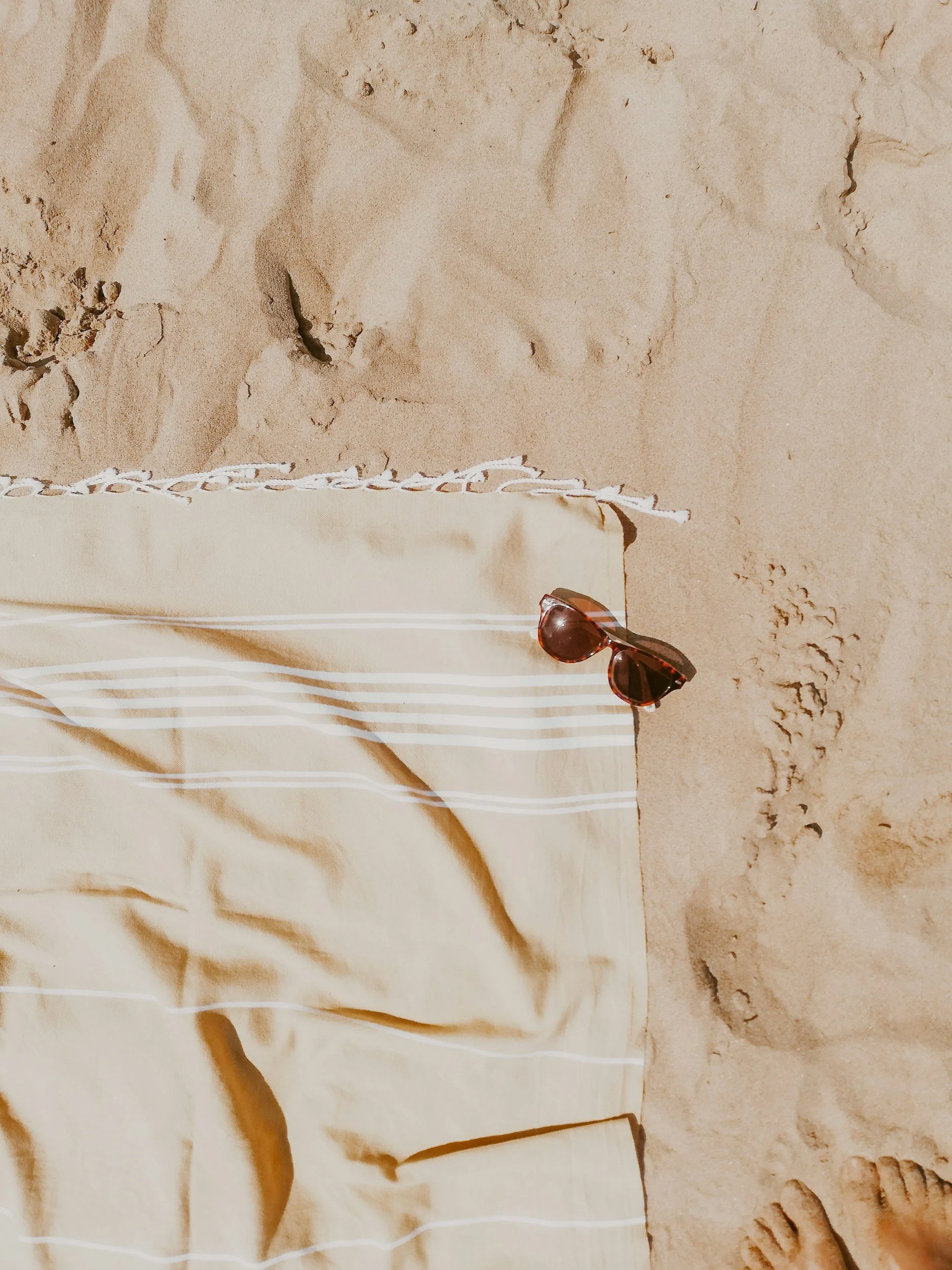 A beige and white striped towel, a pair of sunglasses with dark lenses, and footprints in the sand on a beach indicating a relaxing holiday in the Southwest.