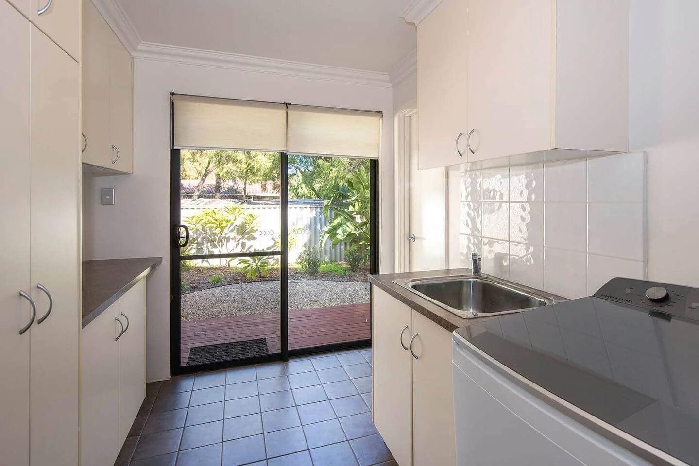Kitchen with white cabinets, a stainless steel sink, a white tiled backsplash, a washing machine, and a glass sliding door leading to an outdoor patio with trees and plants.