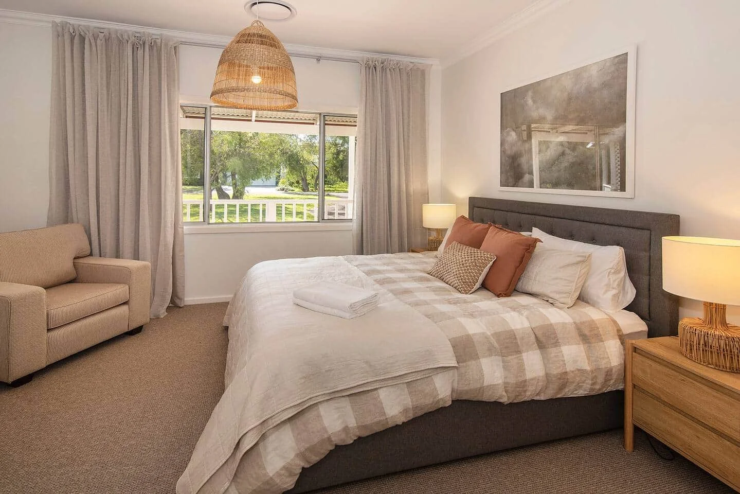 Cozy bedroom with a large bed, beige and white checkered comforter, pillows, wooden nightstand with a lamp, and a window showing trees outside.