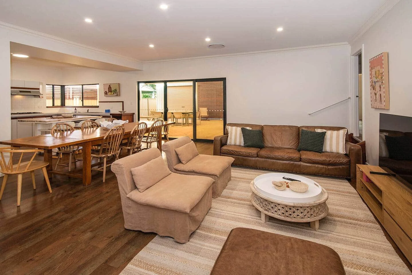 Living room with beige sofas, mixed pillows, wooden coffee table with decorations, area rug, dining table with chairs, sliding glass door leading outside, and a kitchen in background.