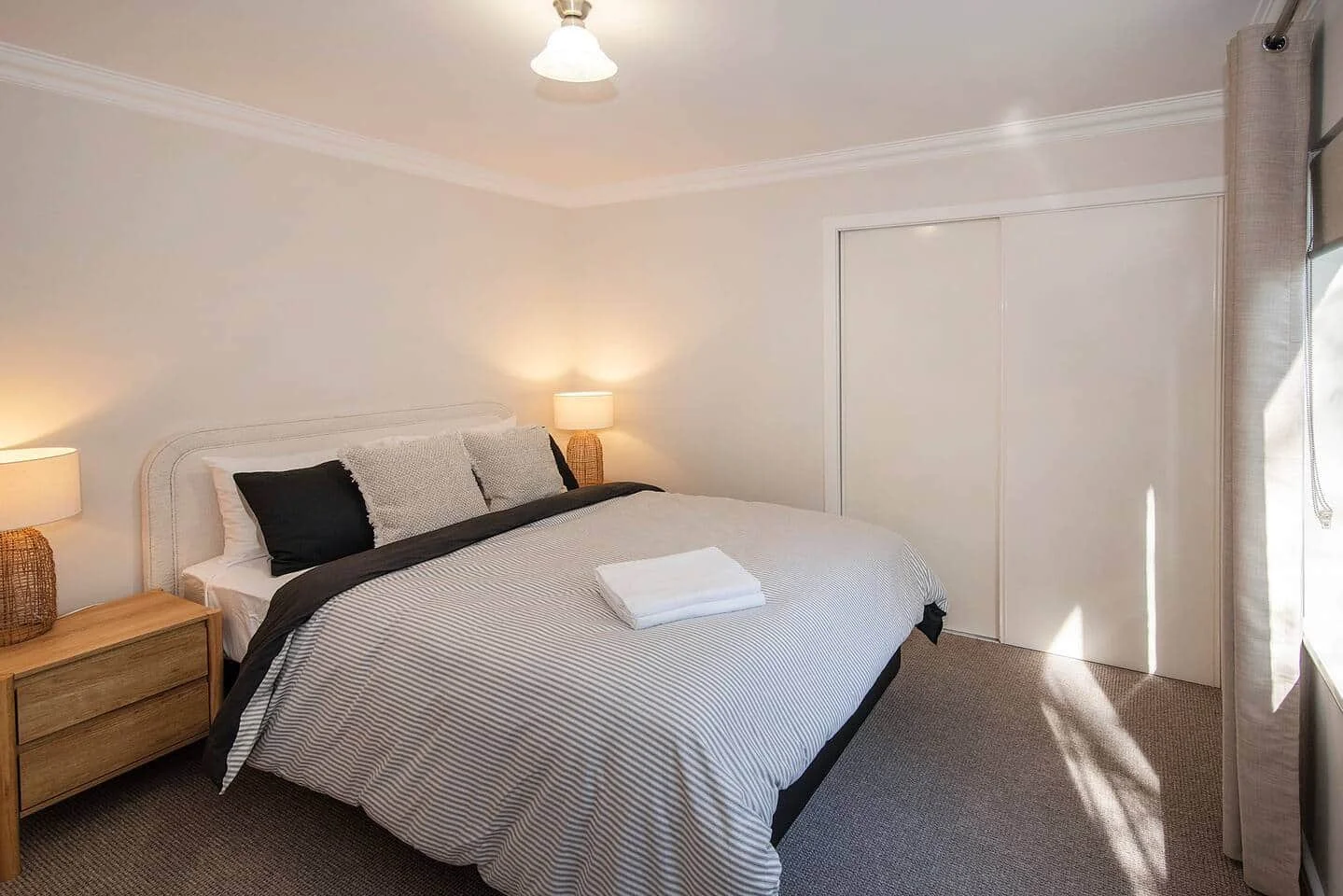 A neatly made bed with striped bedding, black and white pillows, two bedside lamps, a window with curtains, and a closet door in a minimalistic bedroom.