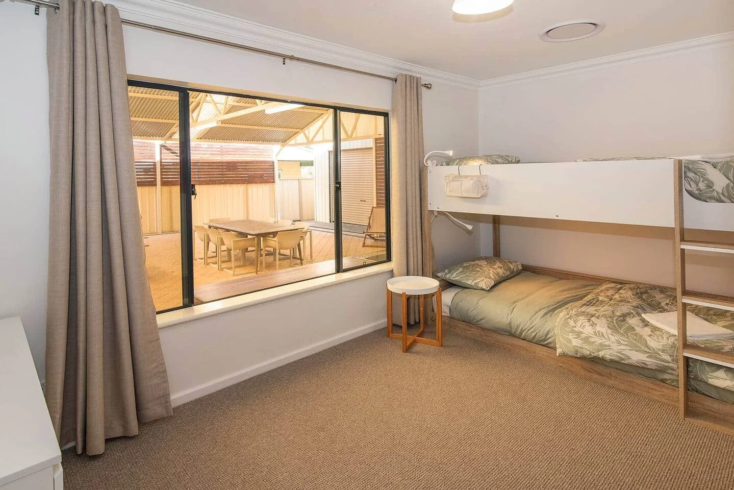 A room with bunk beds for kids or teenagers, a window with curtains, and a small side table. Outside the window, there is a covered outdoor dining area with a table and chairs.