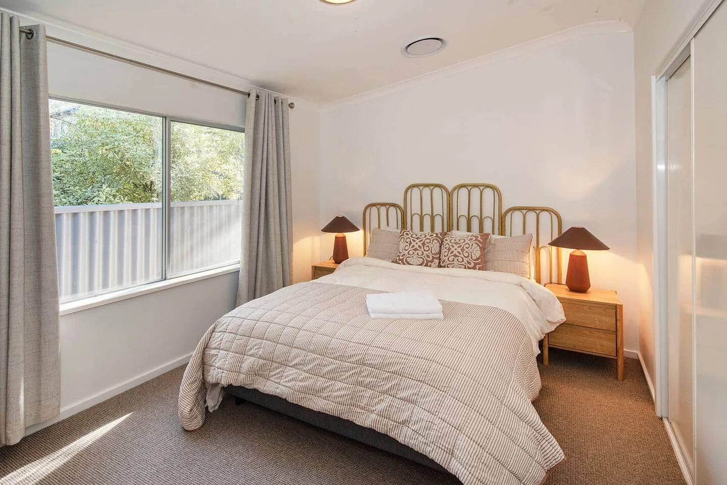 A neatly made bed with a beige striped bedspread, decorated with pillows, flanked by two wooden nightstands with matching lamps. There is a window with open curtains revealing a view of greenery outside.