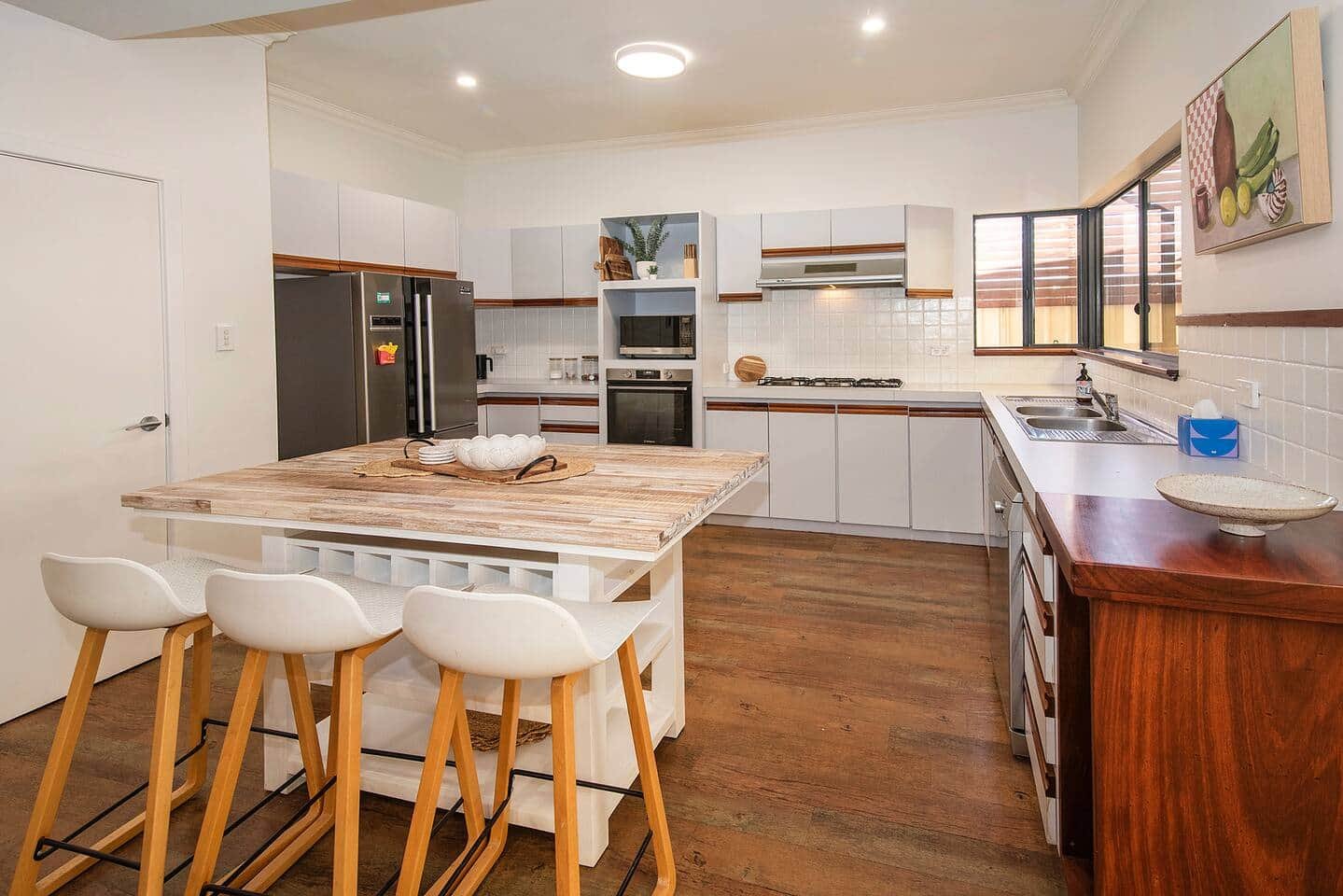 Modern kitchen with white cabinets, wooden accents, a kitchen island with white chairs, stainless steel appliances, and a window above the sink.
