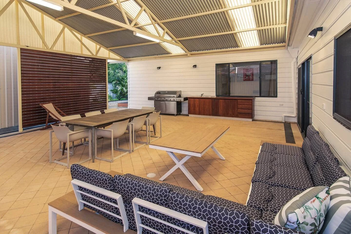 Covered patio with outdoor seating, dining table, BBQ, and outdoor sink, with tiled flooring and white walls.
