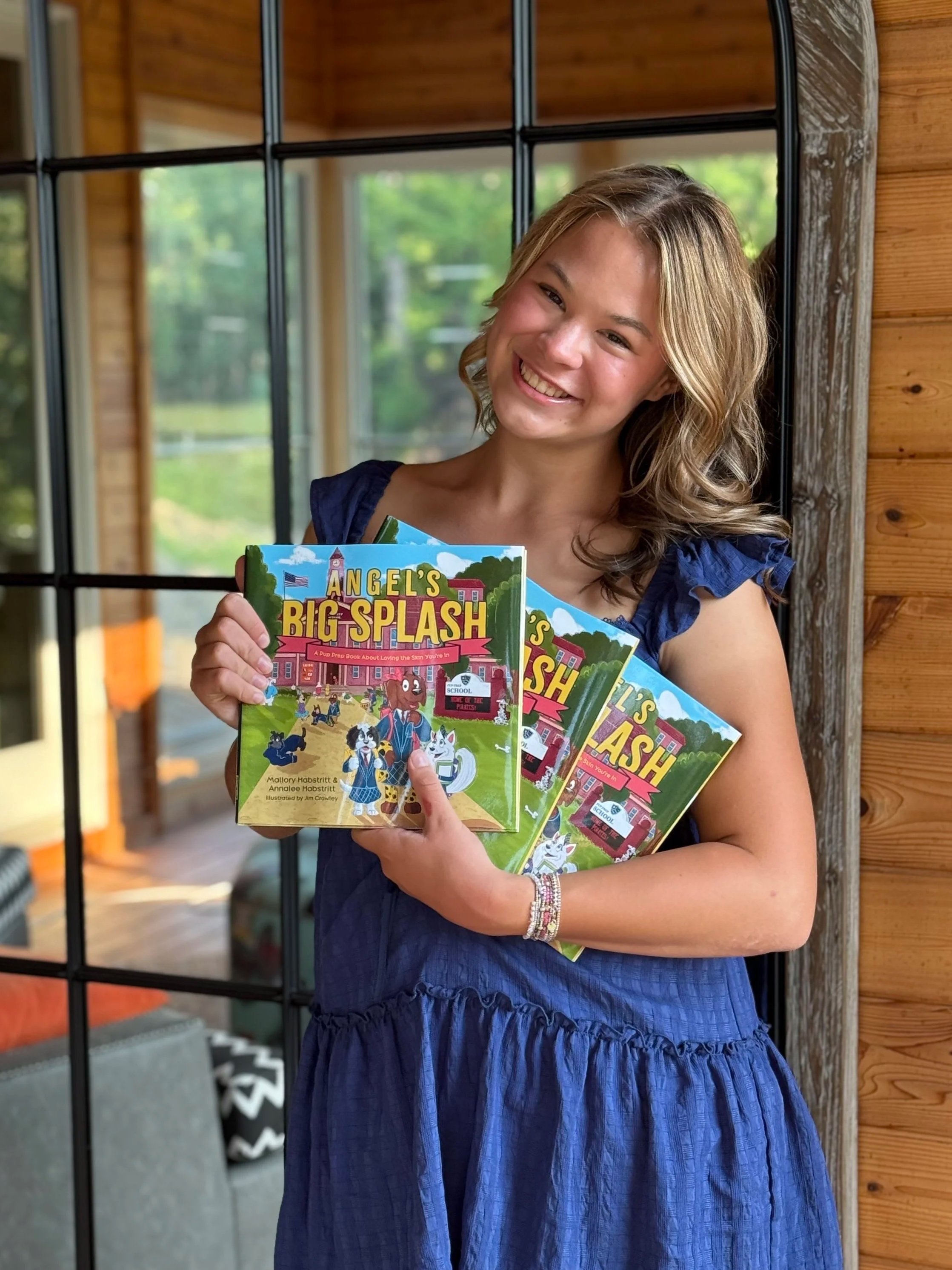 Mallory holding colorful books titled 'Angel's Big Splash: A Pup Prep Book About Loving The Skin You're In' standing indoors near a wooden wall and a window.