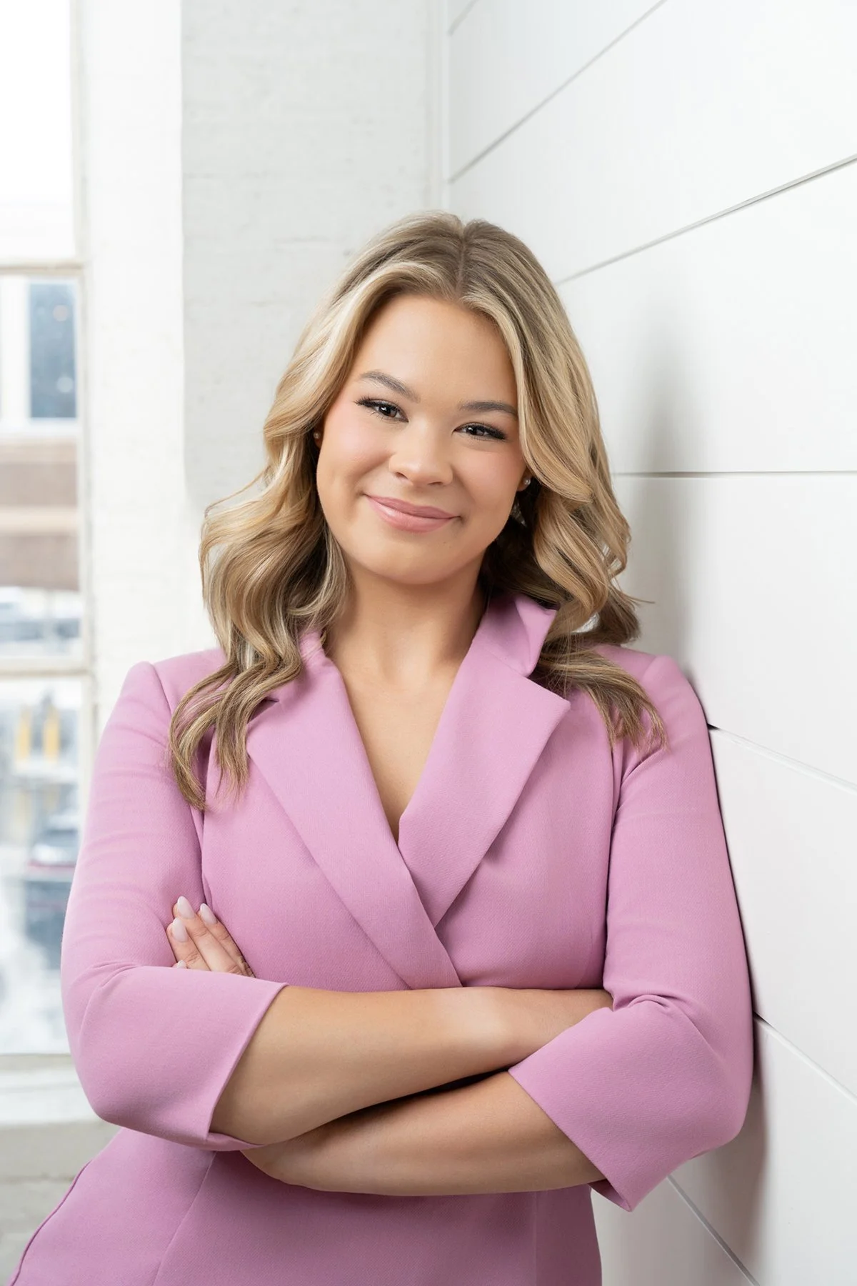 Children's Author, Mallory Habstritt, posing for her headshots. She is smiling wearing a light pink blazer, standing with arms crossed near a white wall in a bright outdoor space.