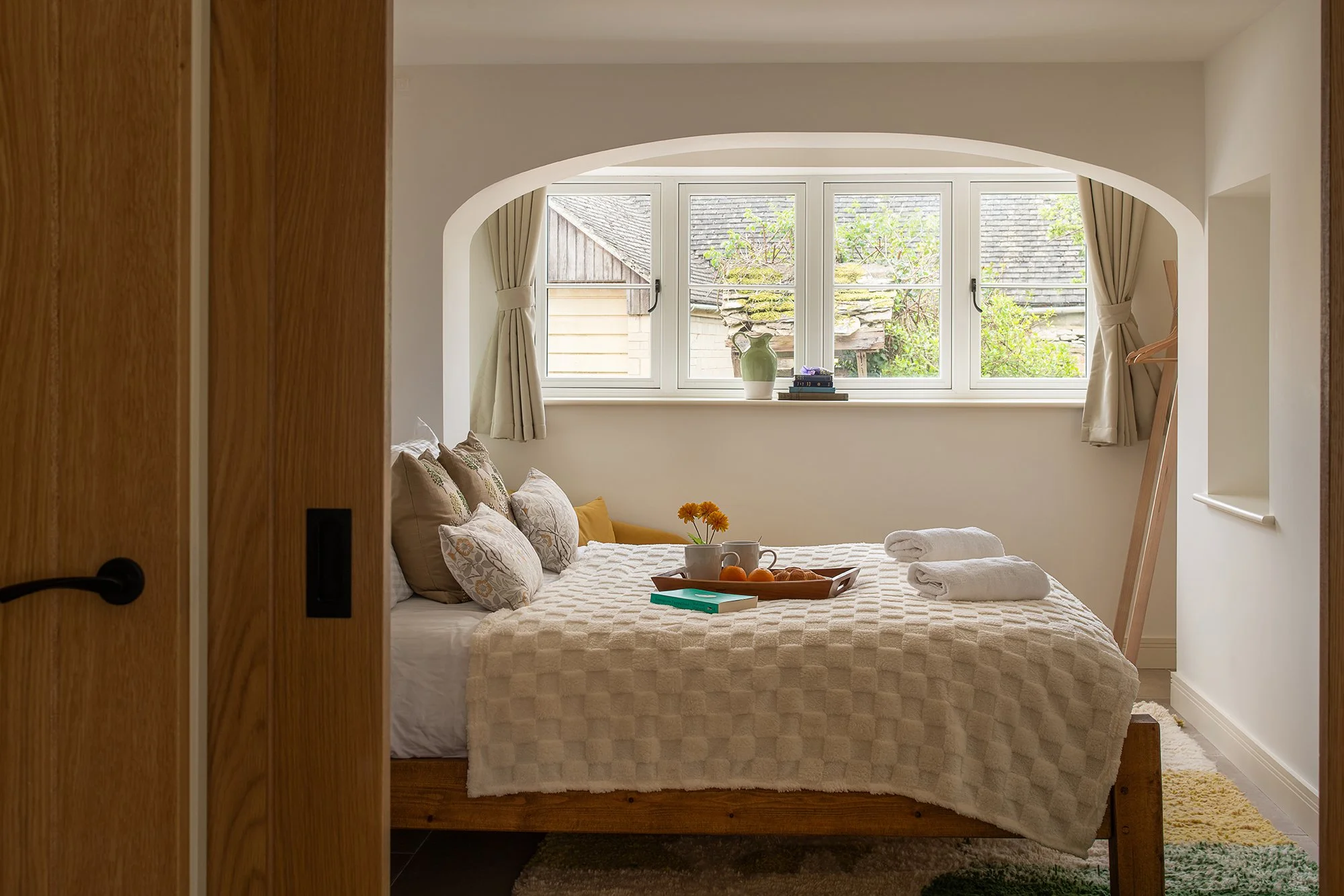 View through an oak door into a cozy holiday cottage bedroom featuring a wooden bed dressed with crisp linens, plush pillows, and a breakfast tray in front of an arched window.