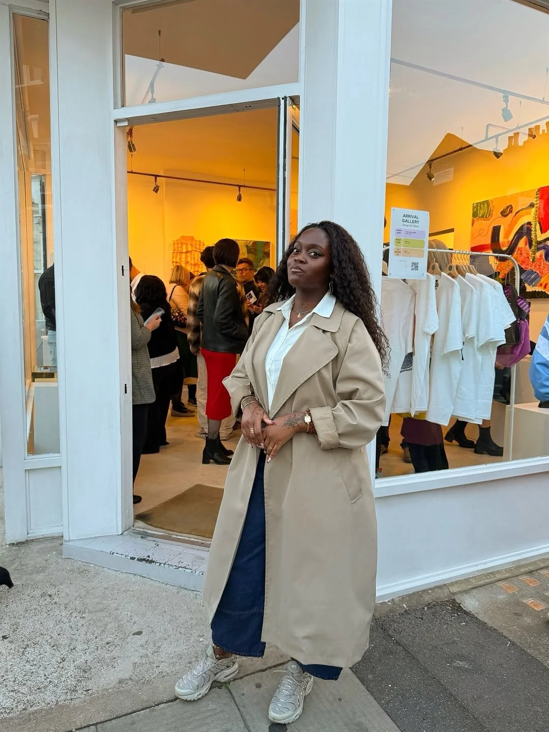 Woman standing outside a gallery entrance, wearing a beige trench coat, white shirt, dark pants, and sneakers, with a crowd visible inside the gallery.