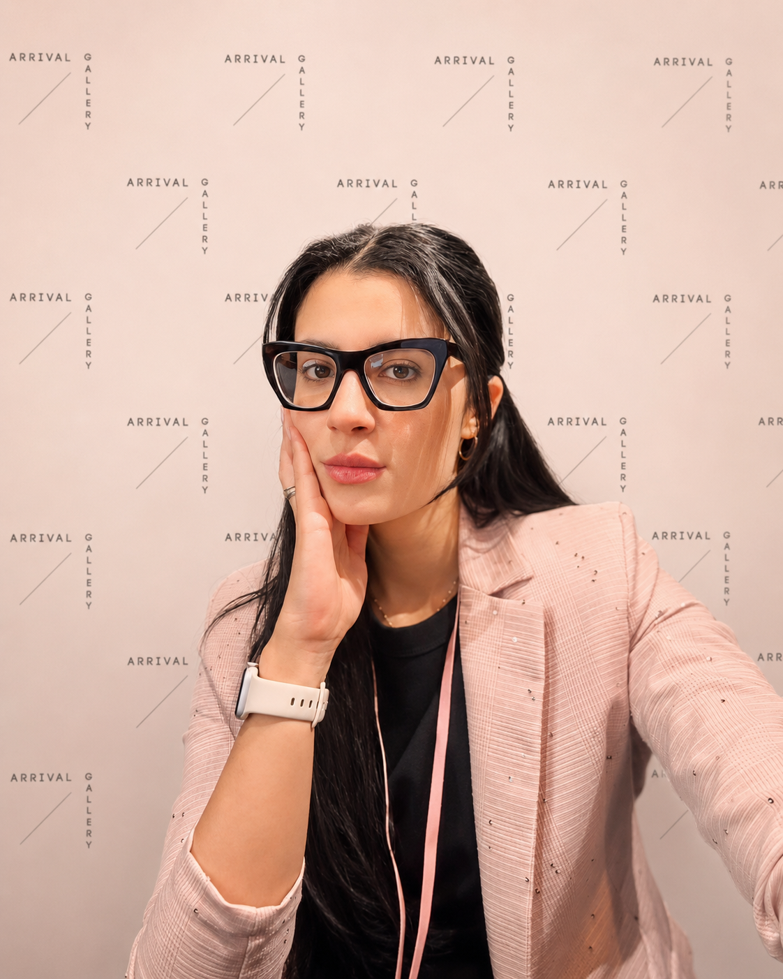 A woman with long dark hair wearing glasses, a pink blazer, and a black shirt, sitting in front of a backdrop that says "Arrival Gallery" repeatedly. She is resting her face on her left hand and looking at the camera.