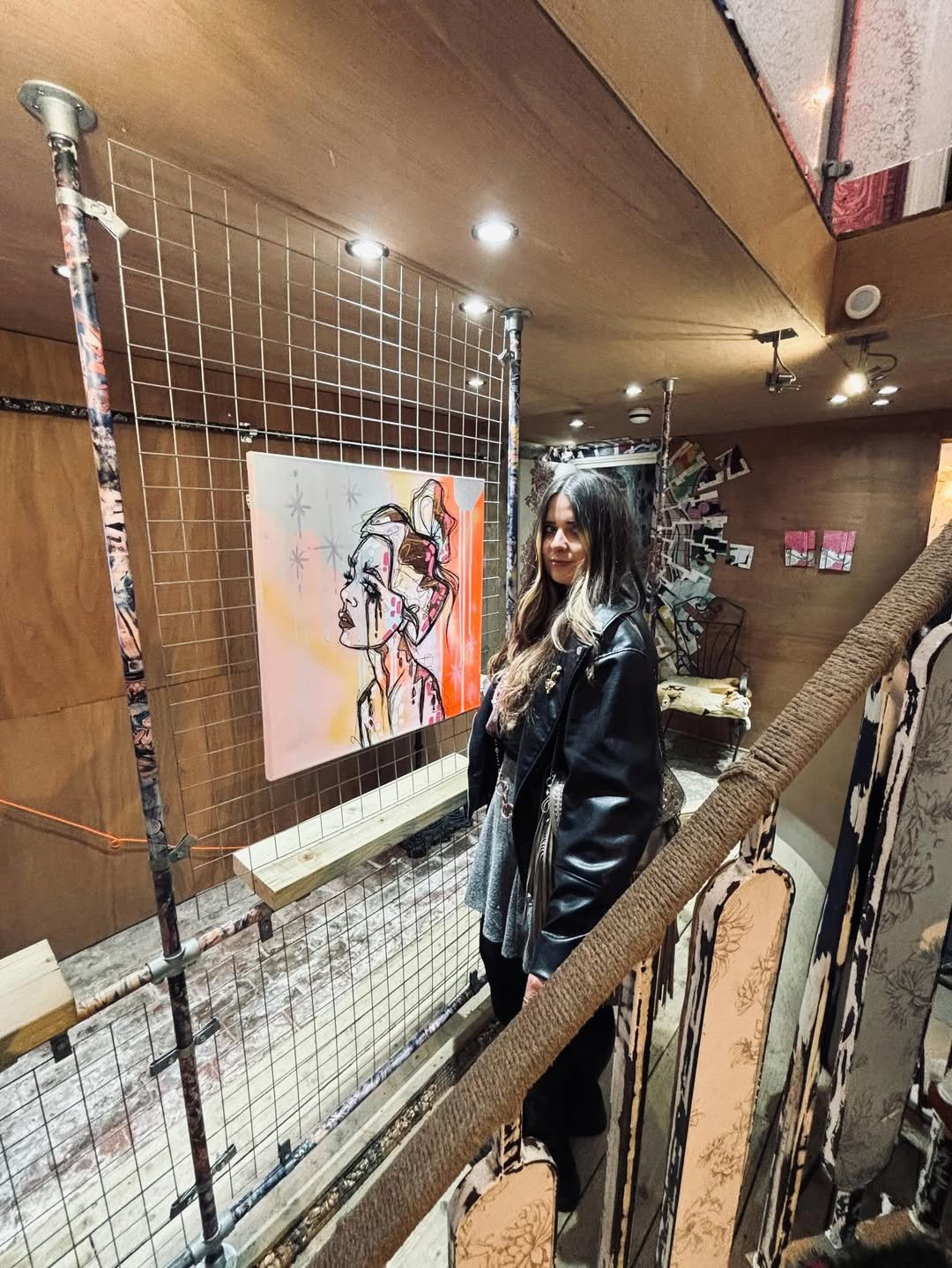 A woman with black and blonde hair wearing a black leather jacket standing in front of a wire grid display with a colorful abstract line art portrait of a woman on it, in an art gallery or studio space.