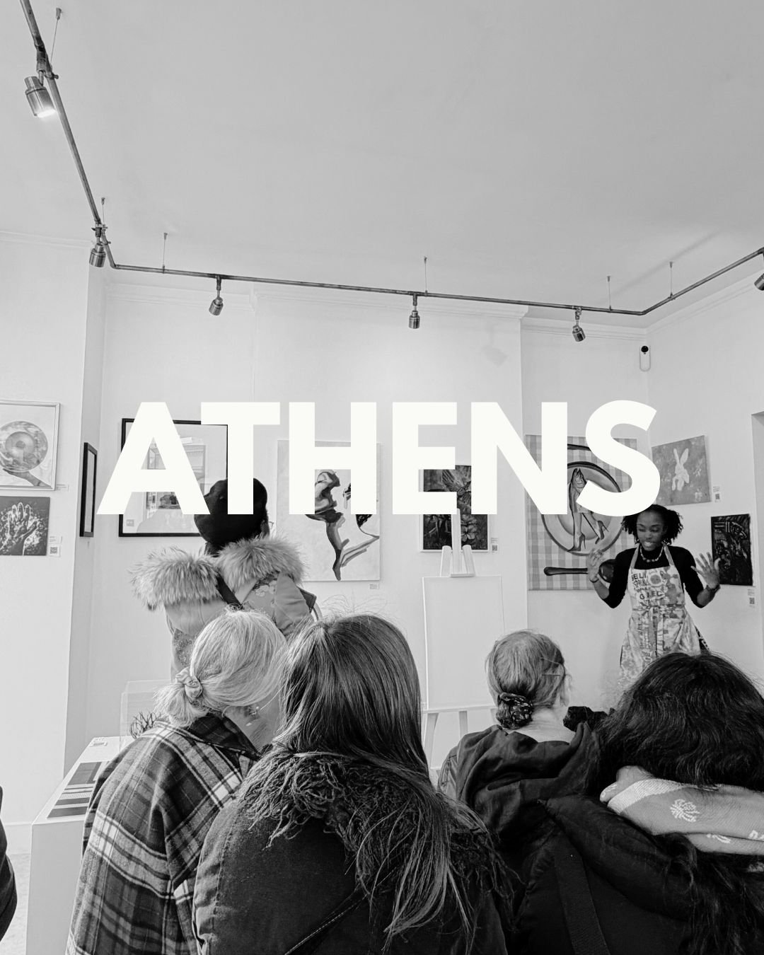 Black and white photo of people attending an art event in Athens, with artwork displayed on the walls and a speaker addressing the audience.
