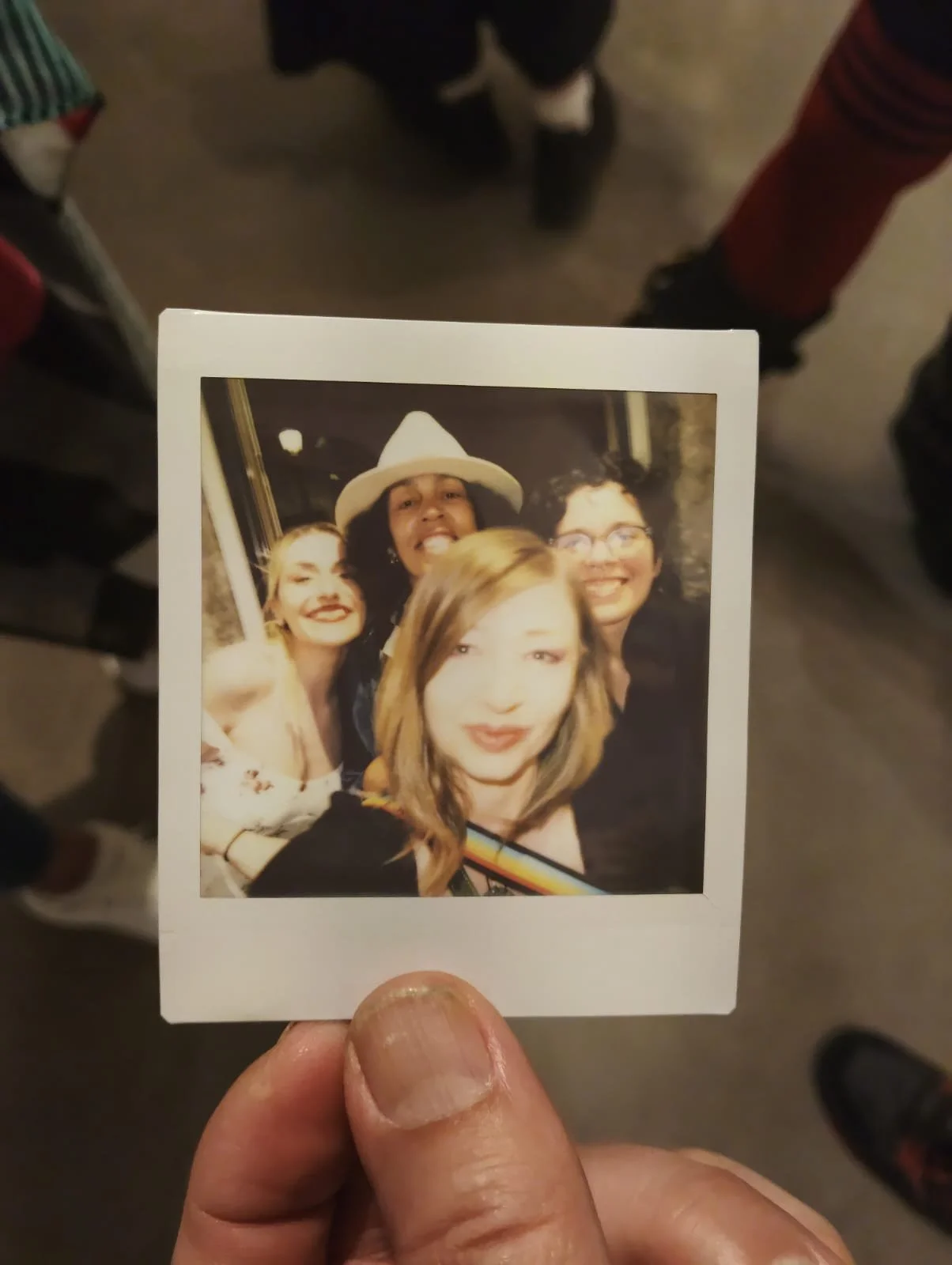 A hand holding a polaroid photo of four smiling friends, one wearing a white hat, in a social setting with other people's legs visible in the background.