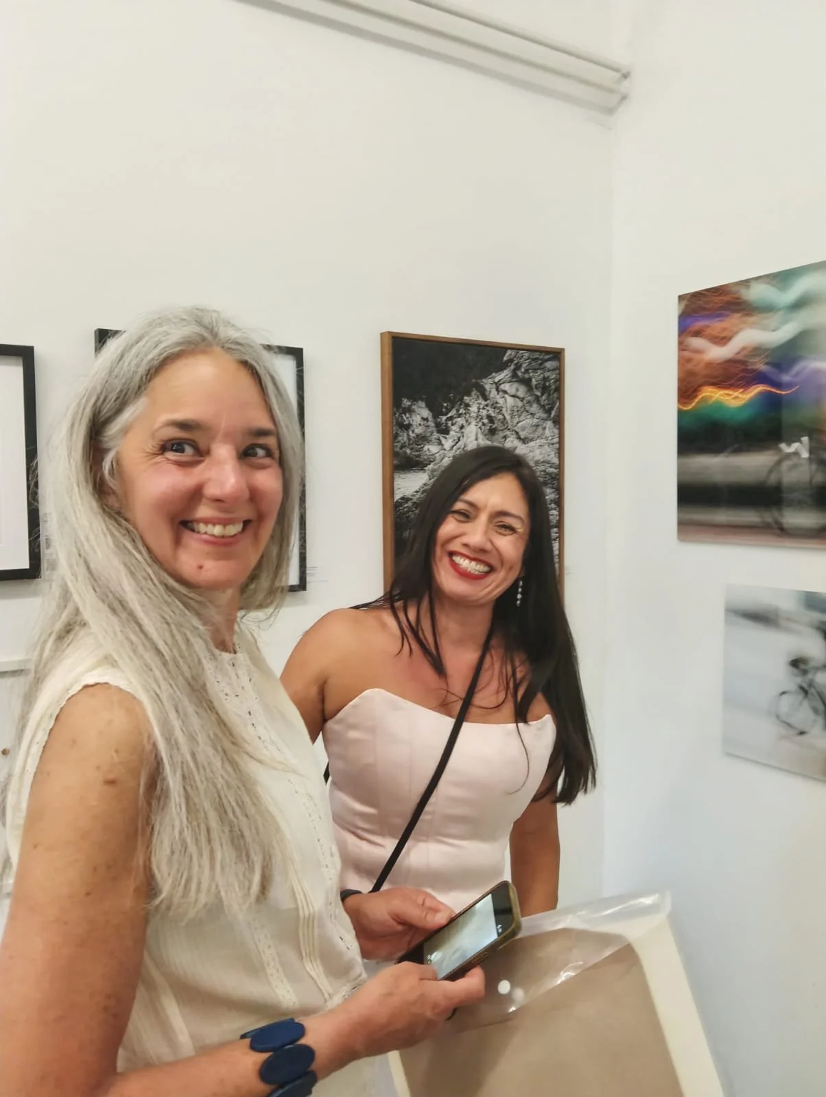 Two women smiling at an art gallery, looking at the camera. One has long gray hair and is holding a smartphone, the other has long dark hair and is wearing a strapless dress, standing in front of framed photographs hanging on a white wall.