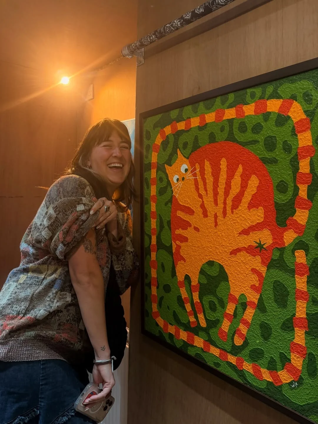 A woman smiling and standing next to a colorful abstract painting of a cat on a wooden wall, with warm lighting overhead.