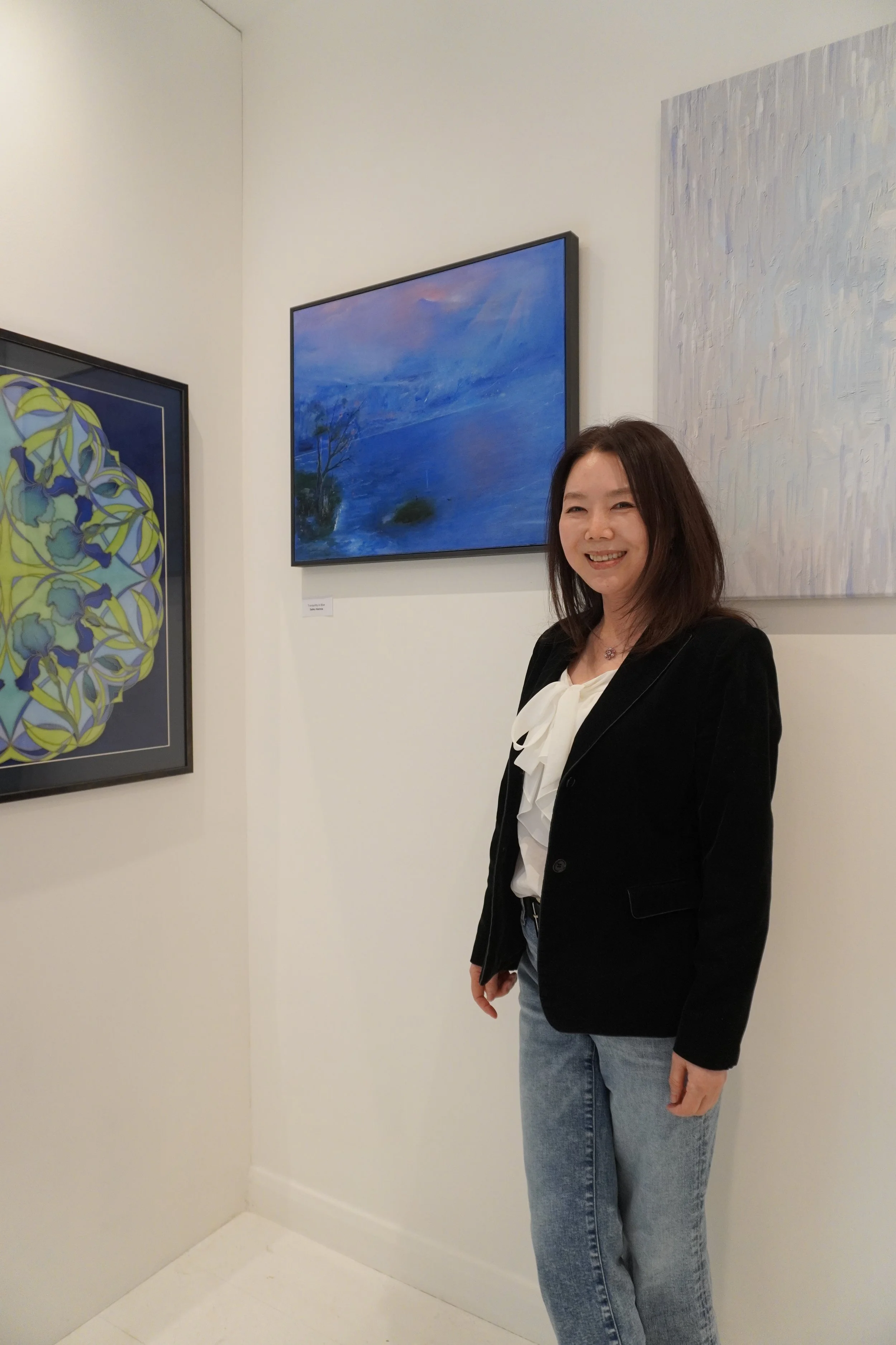 Woman with shoulder-length brown hair smiling at an art gallery, wearing a black blazer, white blouse with a bow, and blue jeans, standing in front of abstract paintings on a white wall.