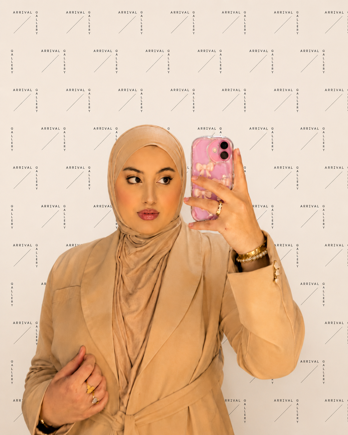 Woman wearing a beige hijab and blazer taking a selfie with a pink phone case featuring bows, against a white backdrop with 'Arrival Gallery' repeated in black text.