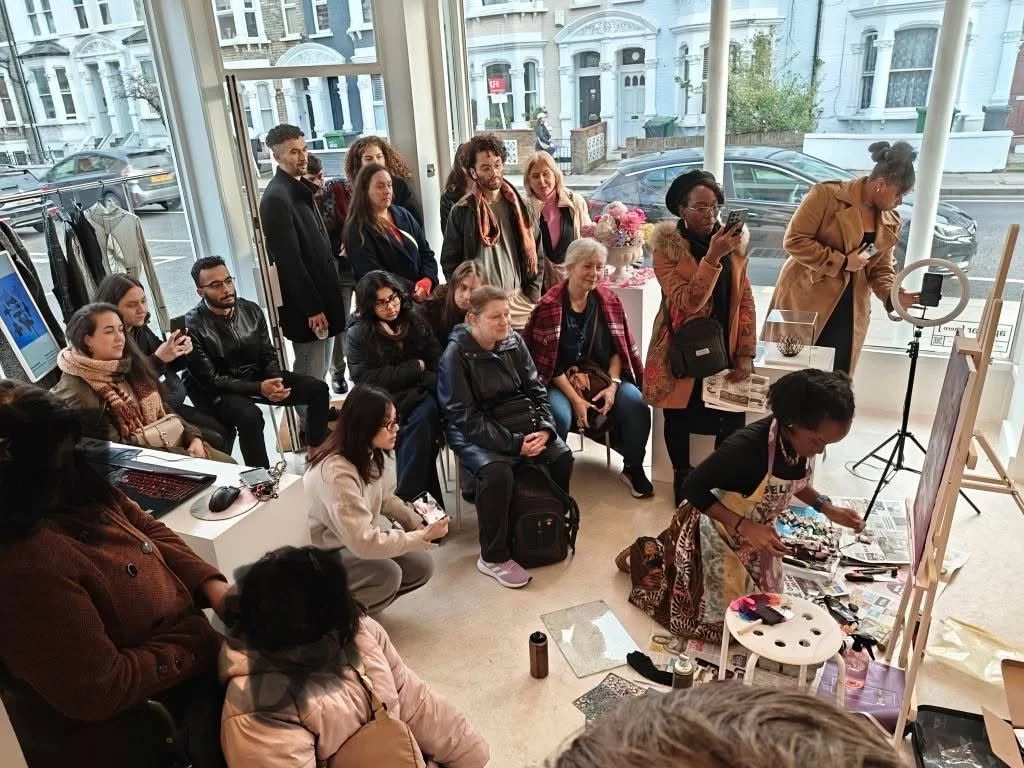 Group of people observing an artist creating jewelry in a bright showroom with large windows, and a street scene outside.