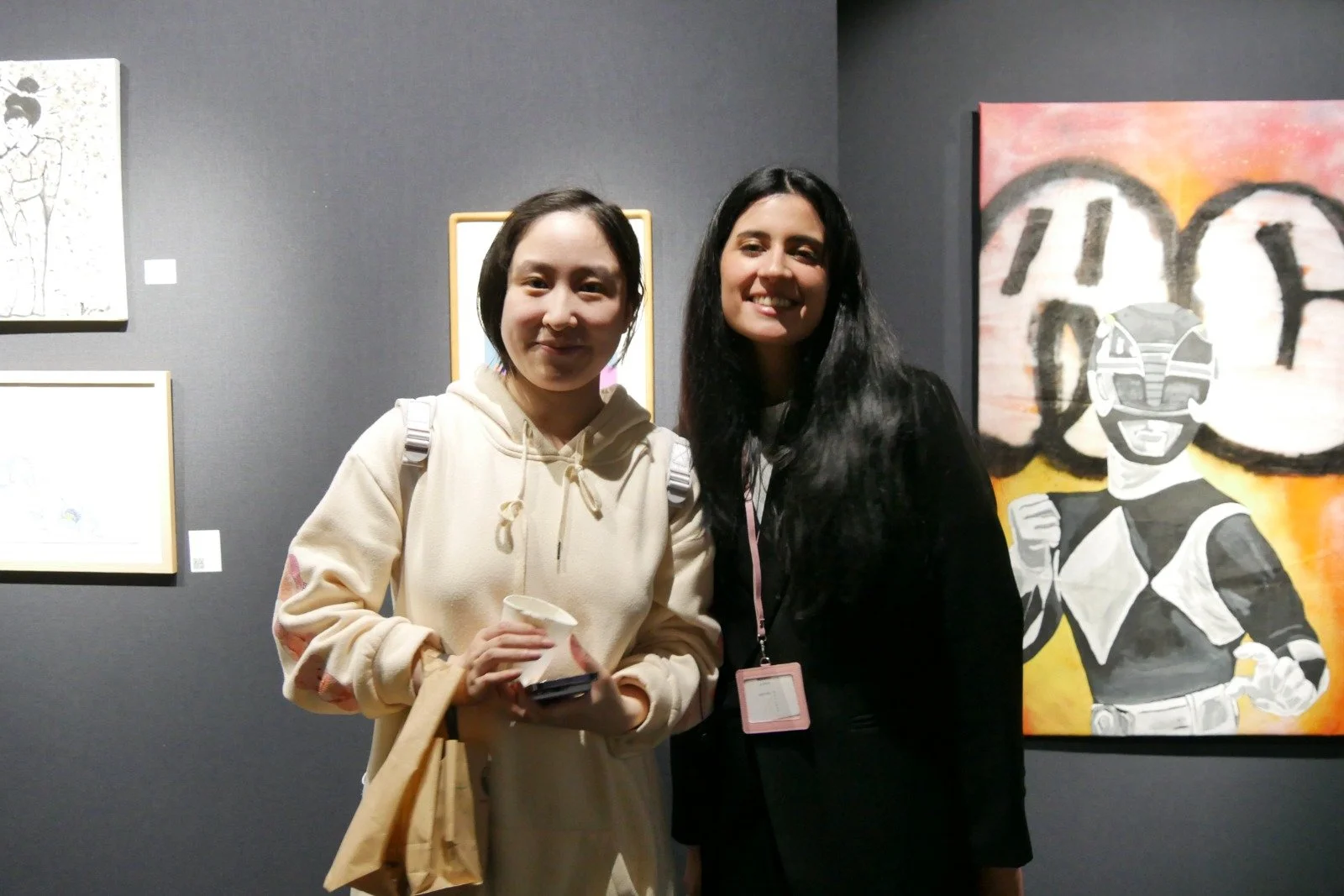 Two women standing in front of artwork at a gallery. One woman is wearing a beige hoodie, holding a paper cup and a phone. The other woman is in black with an ID badge, smiling. The artwork behind features a stylized figure and graffiti-style text.