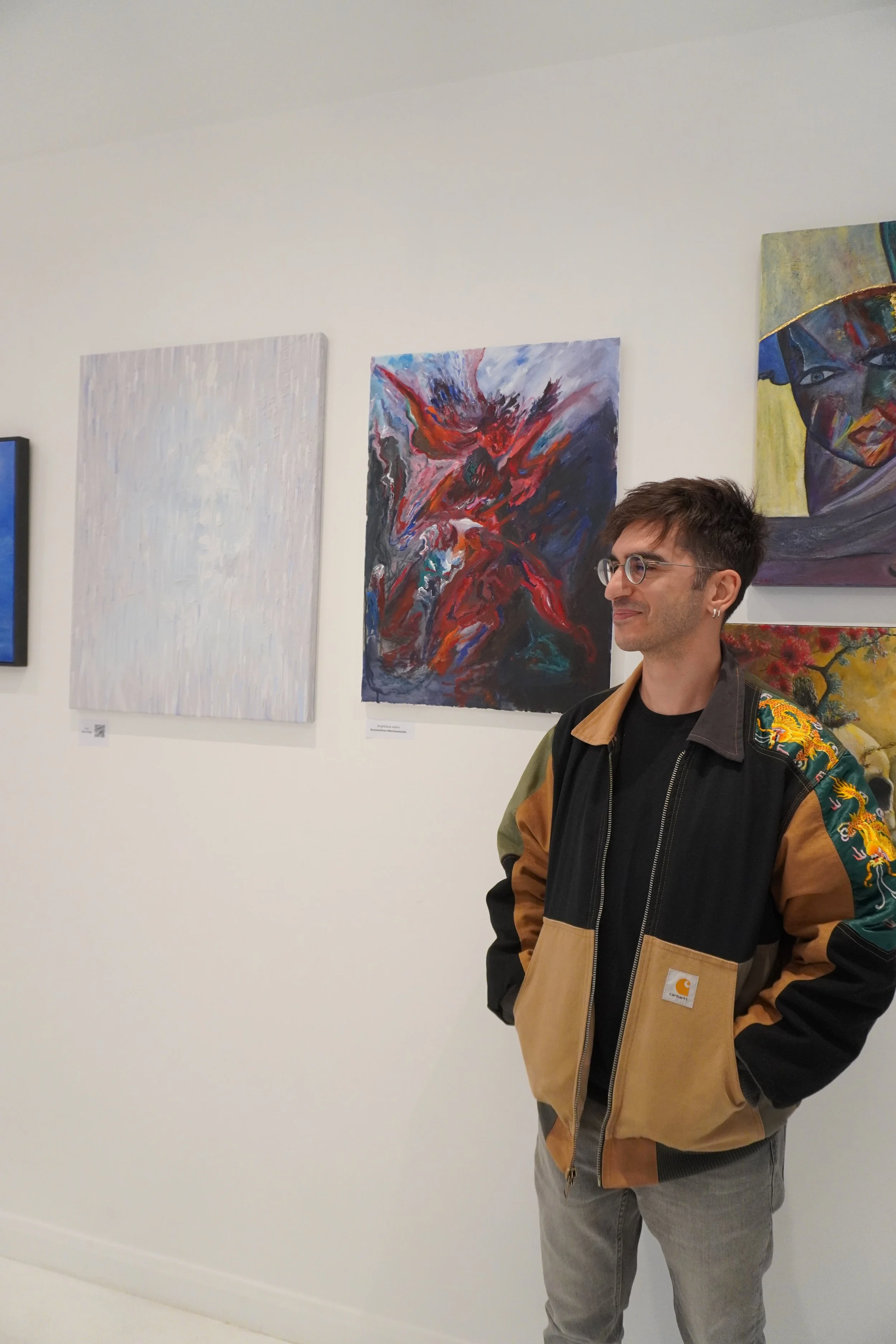 A man with glasses and a small earring standing in front of colorful abstract paintings in an art gallery. He is wearing a Carhartt jacket with a floral pattern on the sleeves and relaxed gray jeans.
