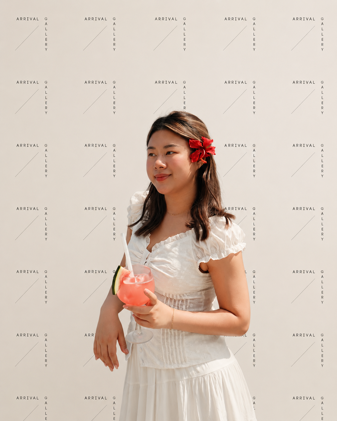 A woman with a red flower in her hair, wearing a white dress, holding a pink cocktail garnished with a lime wedge, standing against a white backdrop with repeated 'Arrival Gallery' text.