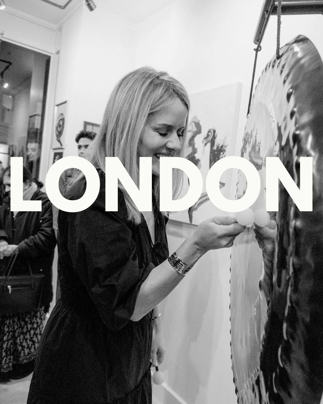 A woman at an art gallery in London is engaging with a piece of art on display, smiling and holding a cotton swab, while other people are visible in the background.