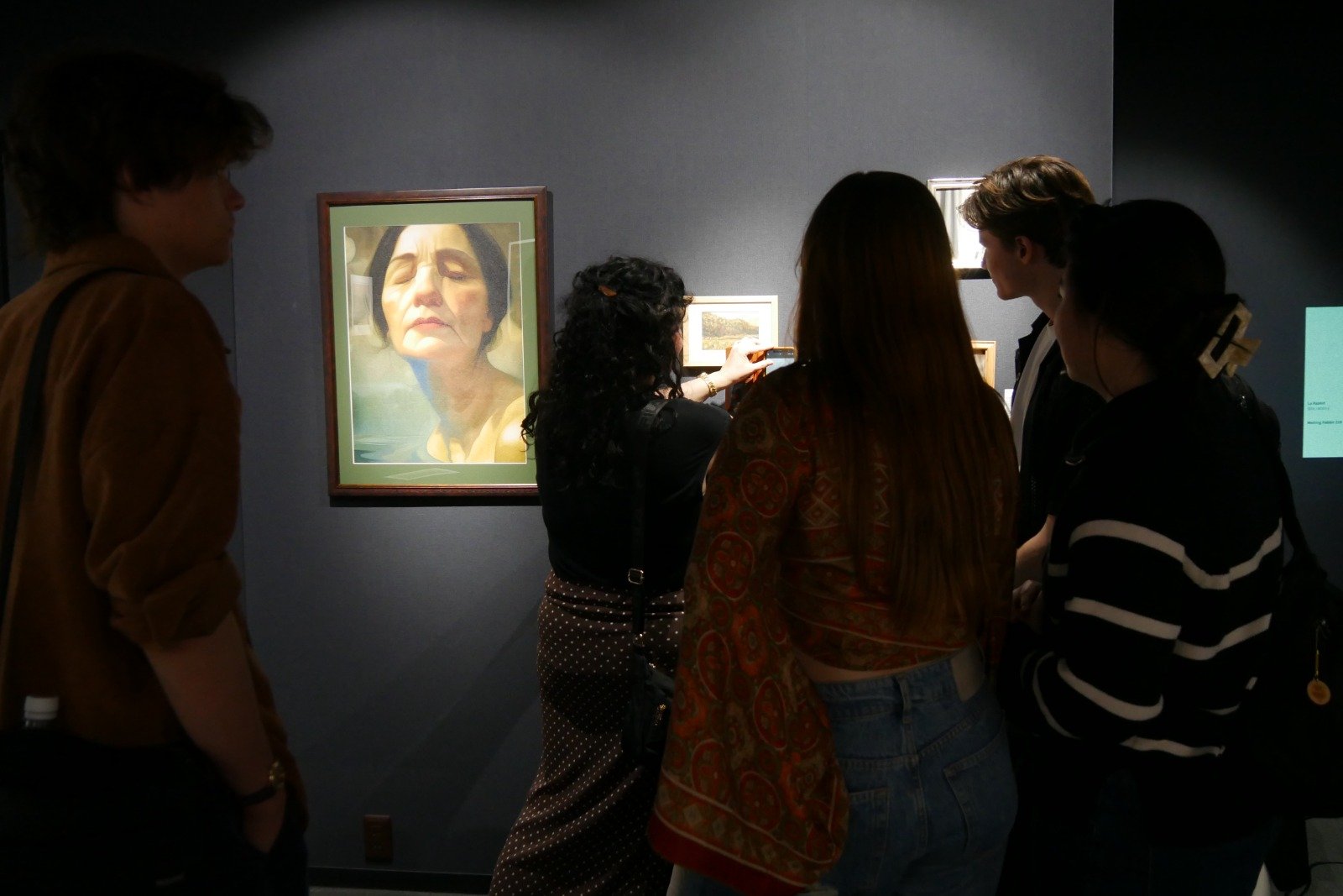 A group of people viewing artwork in a gallery, including a portrait of an older woman with short dark hair and closed eyes.
