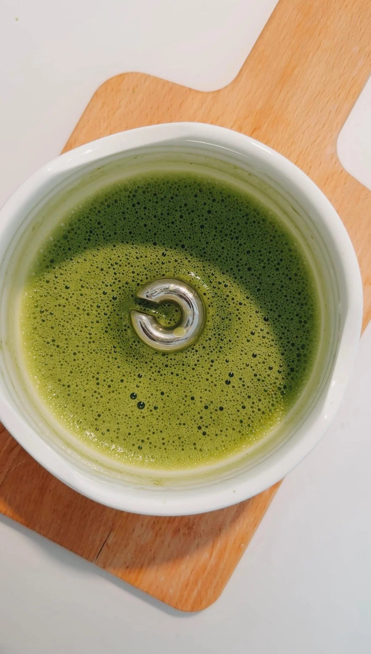 A top-down view of a bowl of green matcha tea blended with water, placed on a wooden cutting board. With a chunky silver earring or hoop floating aesthetically.