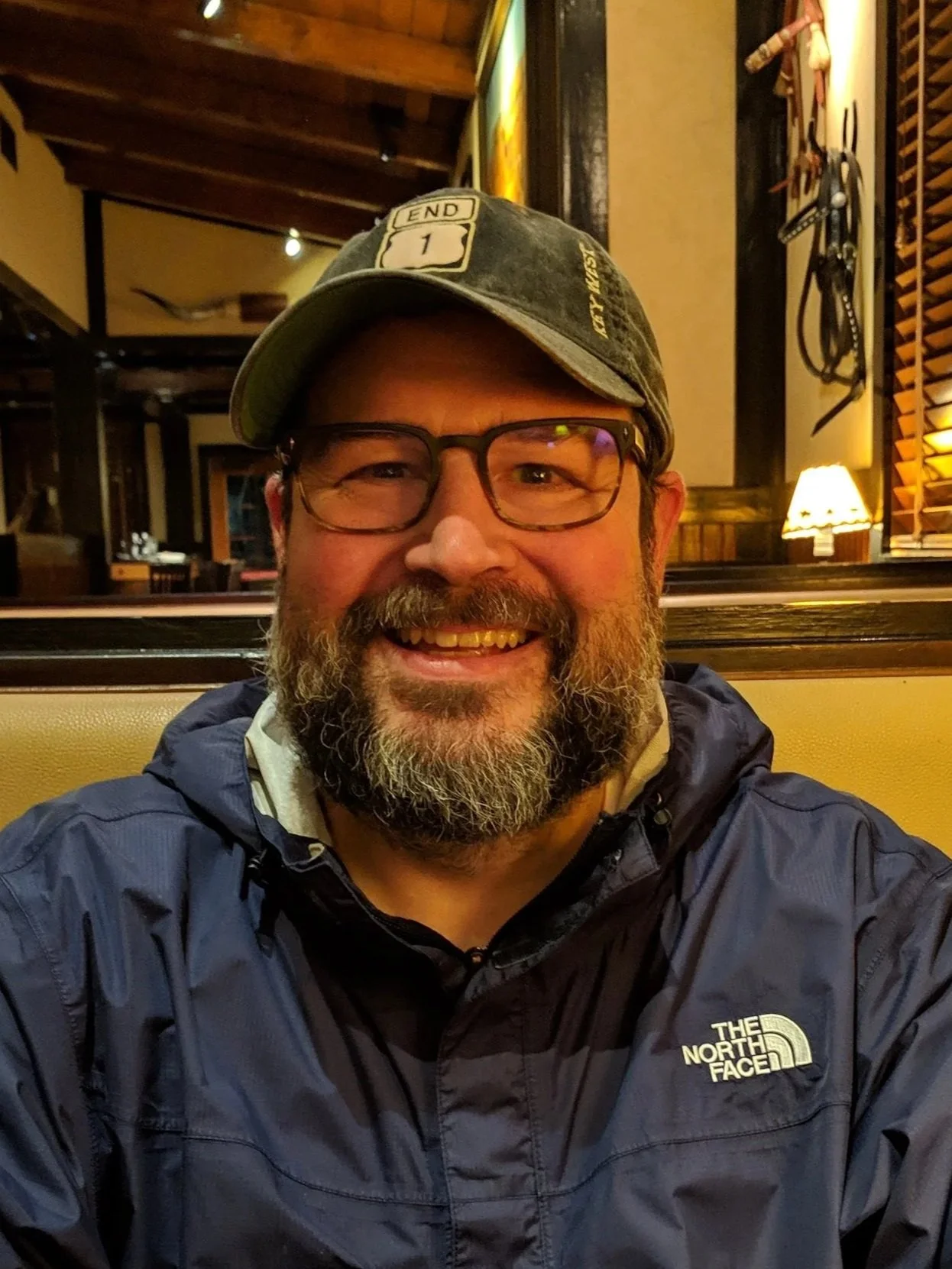 A man with glasses, a beard, and a baseball cap smiling at the camera, wearing a blue The North Face jacket inside a warmly lit restaurant with wooden decor and mounted tools on the wall.