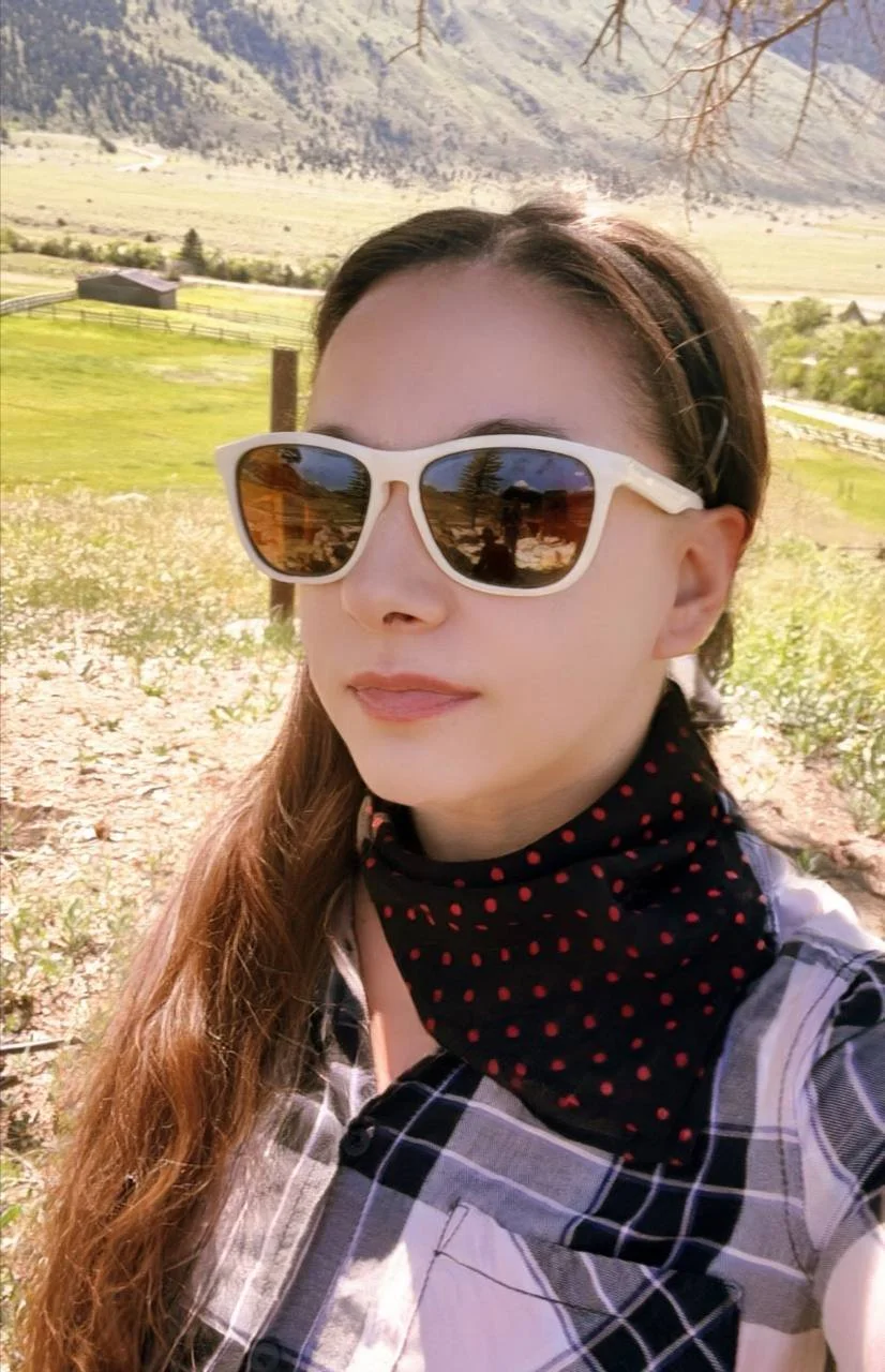 A woman with long brown hair wearing white sunglasses with brown lenses, a black headband, a black and red polka dot neck gaiter, and a gray plaid shirt, standing outdoors with a green field, trees, and mountains in the background.