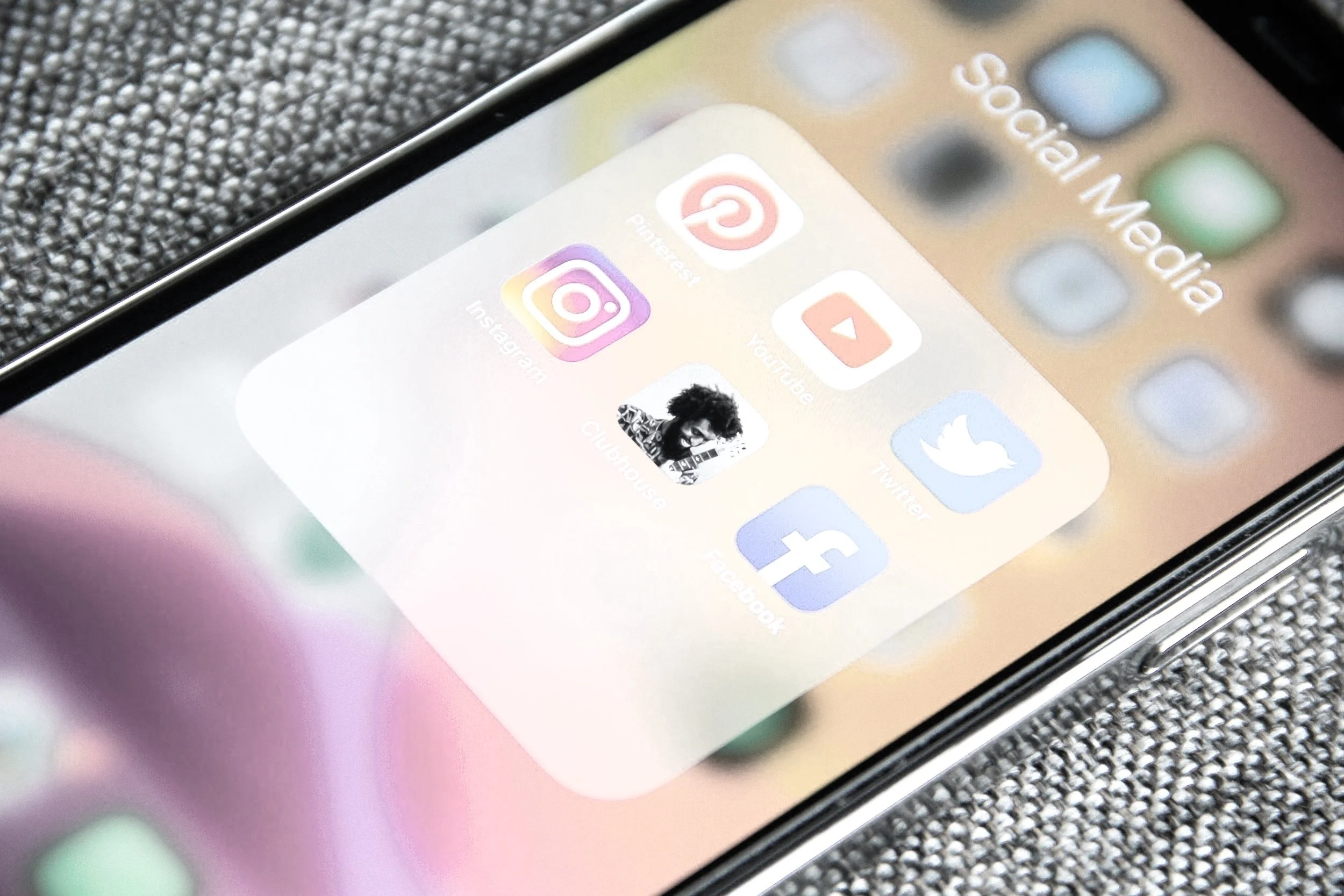 Close-up of a smartphone screen displaying a folder labeled 'Social Media' with apps such as Pinterest, YouTube, Instagram, Twitter, Facebook, and Clubhouse, resting on a textured surface.