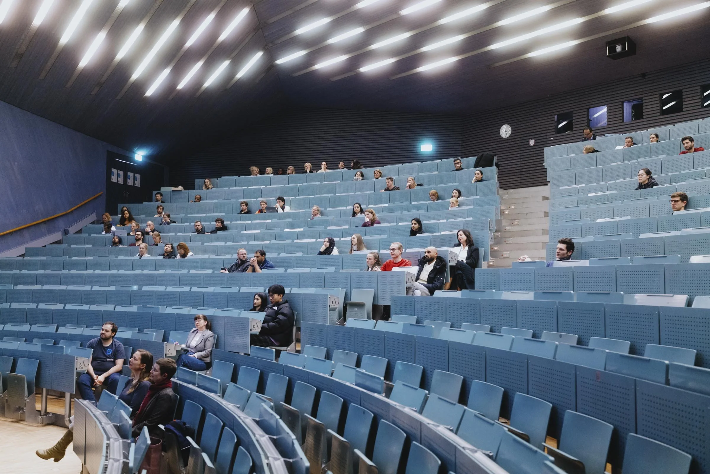 Menschen sitzen in einem großen, modernen Hörsaal mit blauen Stühlen, der an einem Vortrag teilnimmt.
