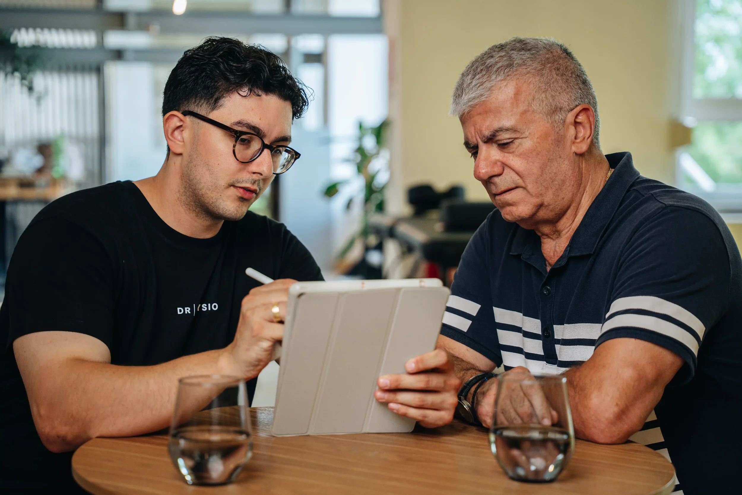 Zwei Männer sitzen an einem Tisch, schauen auf ein Tablet, während sie miteinander sprechen.