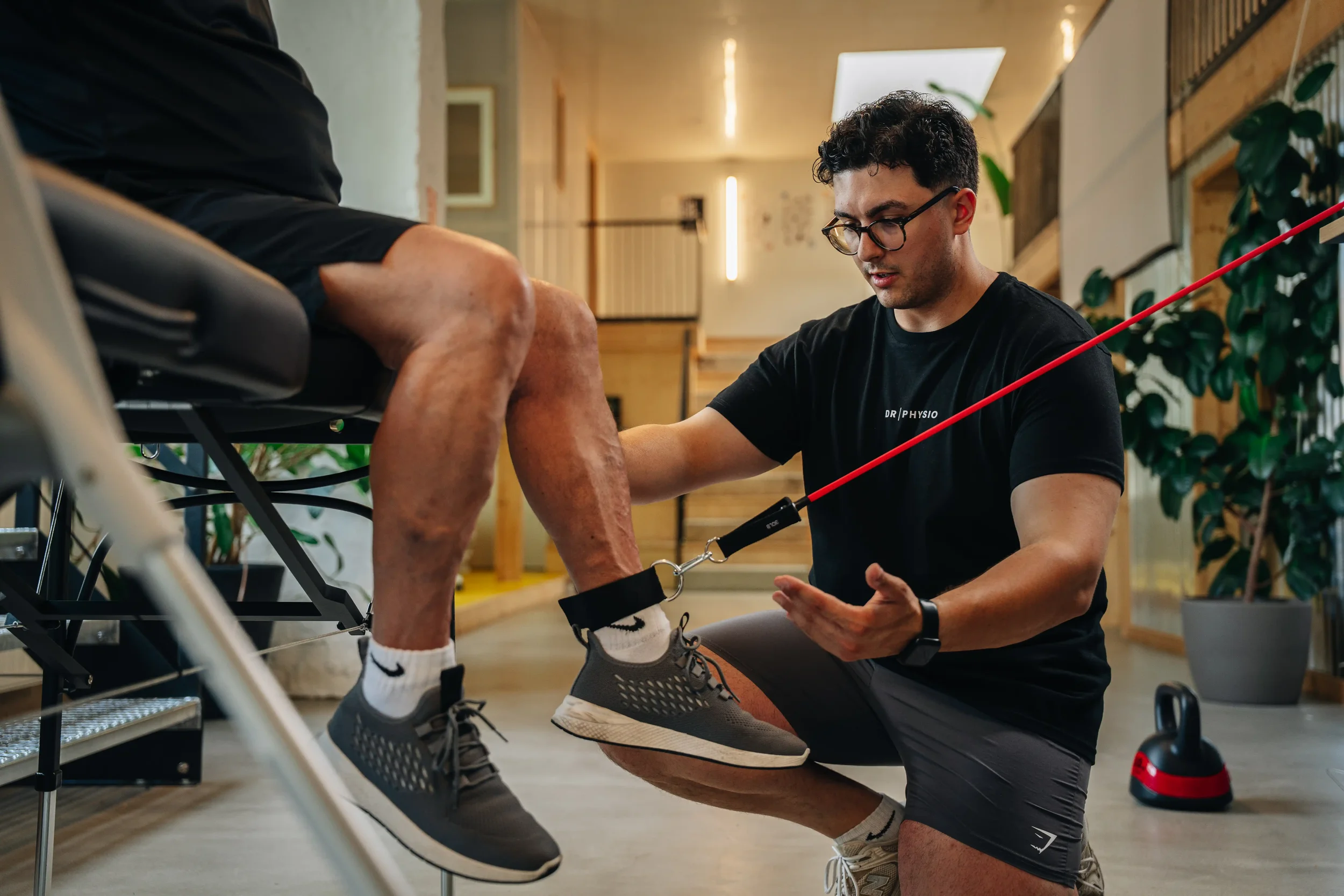 Ein Physiotherapeut trainiert einen Patienten mit einem Trainingstool an einem Bein, im Raum mit Pflanzen und Holzdekor.
