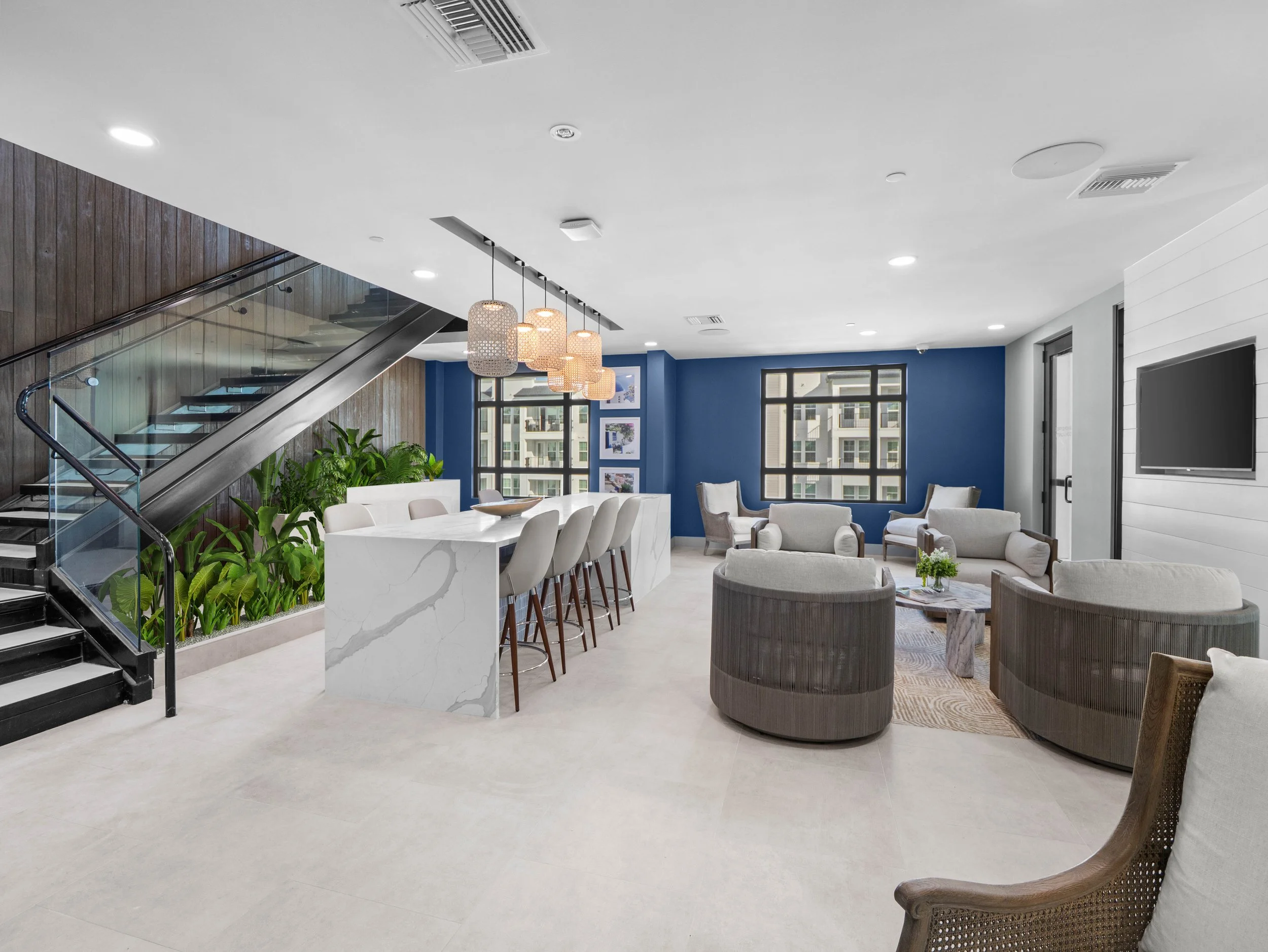 Modern apartment clubhouse lounge with marble kitchen island, bar seating, contemporary pendant lights, staircase, plants, and gray seating area