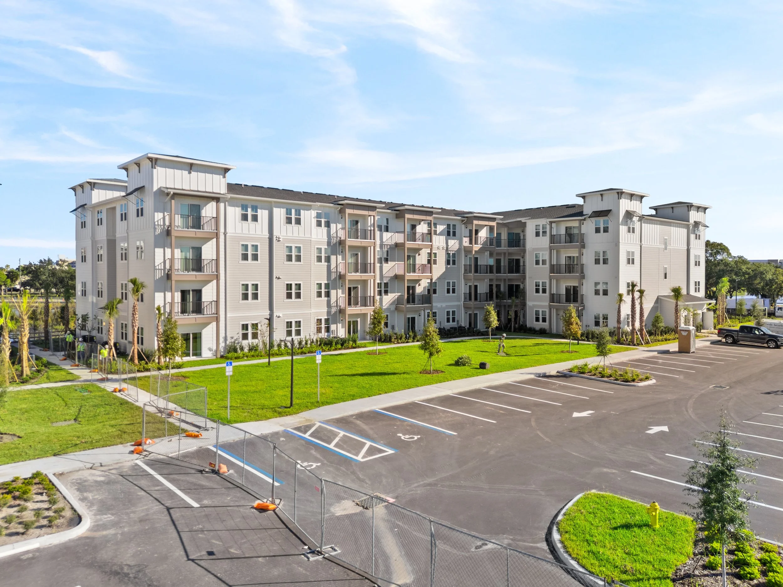 Aerial view of a modern multi-story apartment complex with balconies, landscaped lawn, palm trees, and accessible parking spaces.