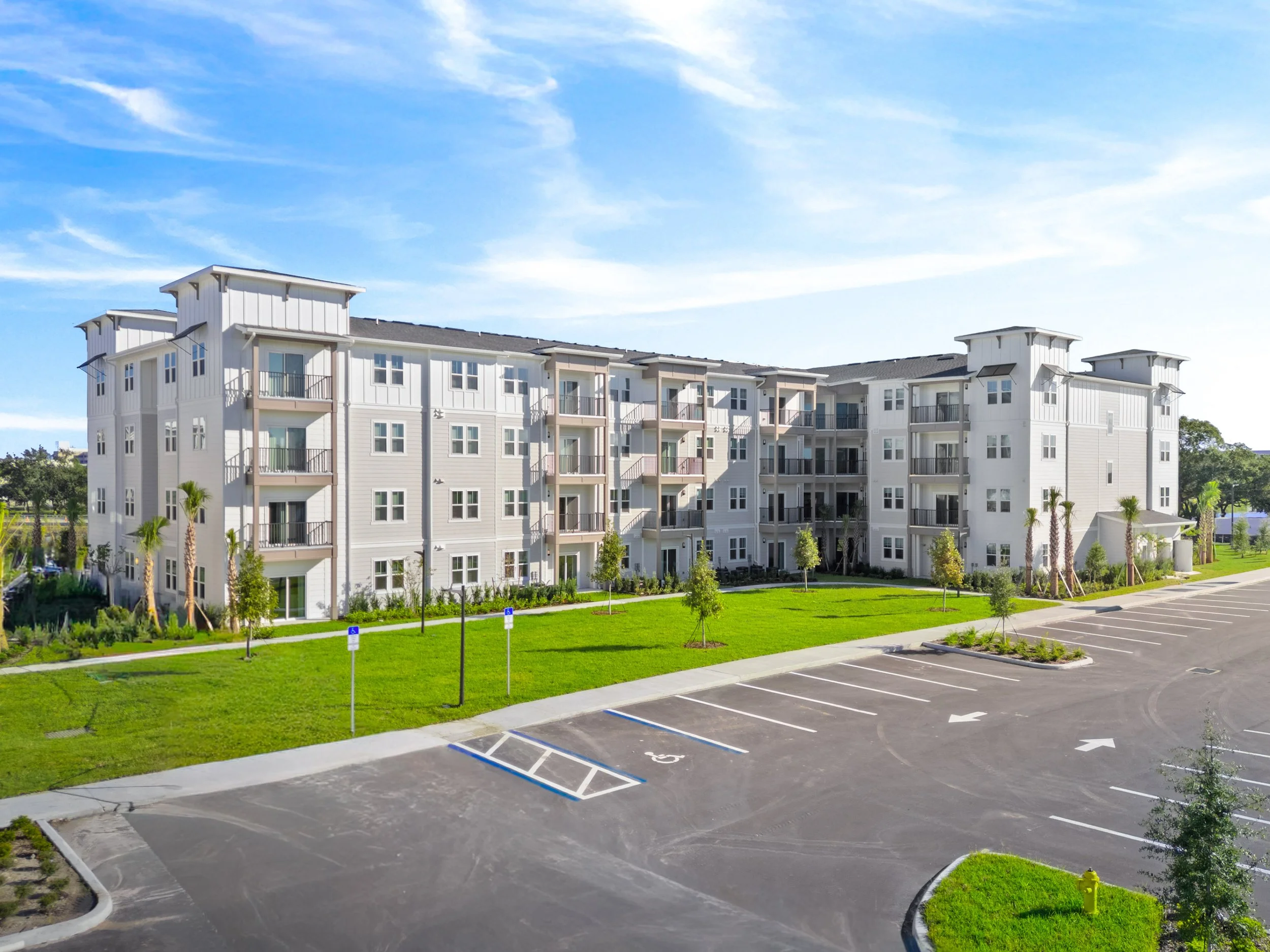 Modern multi-story apartment building with balconies, landscaped lawn, palm trees, and a large parking lot under a blue sky