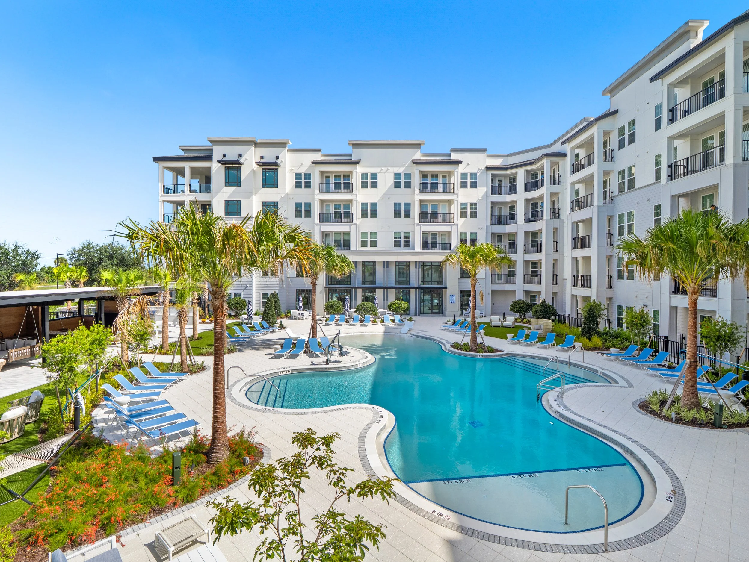 Modern white apartment complex surrounding a resort-style swimming pool with palm trees and blue lounge chairs